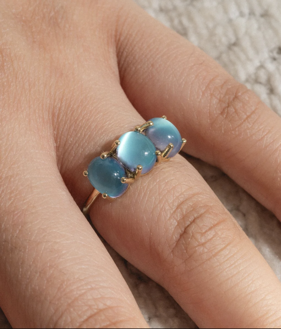 A hand wearing a gold ring set with three oval blue gemstones, displayed against a textured light surface.