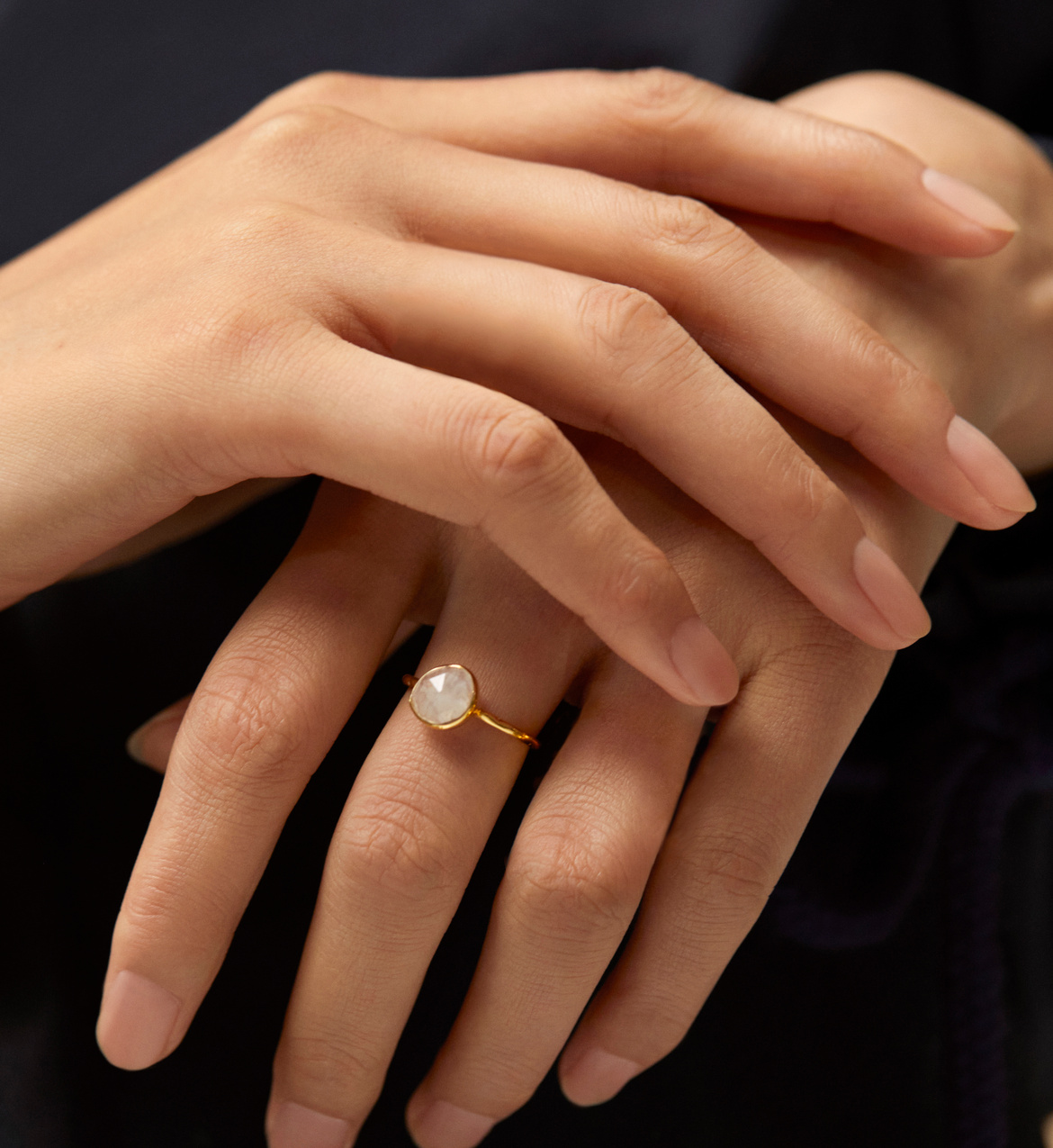 A close-up of two hands, one wearing a gold ring with a round, translucent stone, against a dark background.