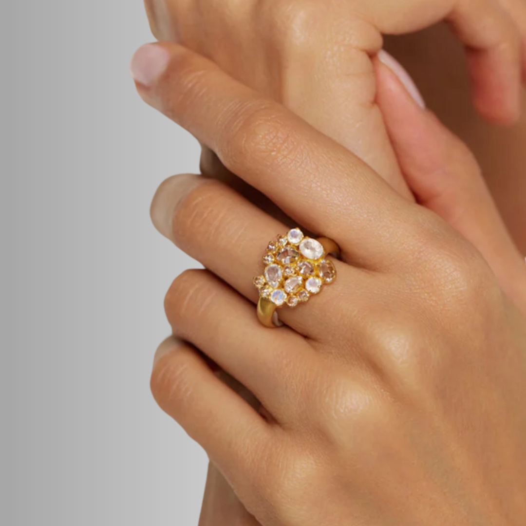 A hand wearing a gold ring with multiple clear and amber gemstones, set against a neutral gray background.