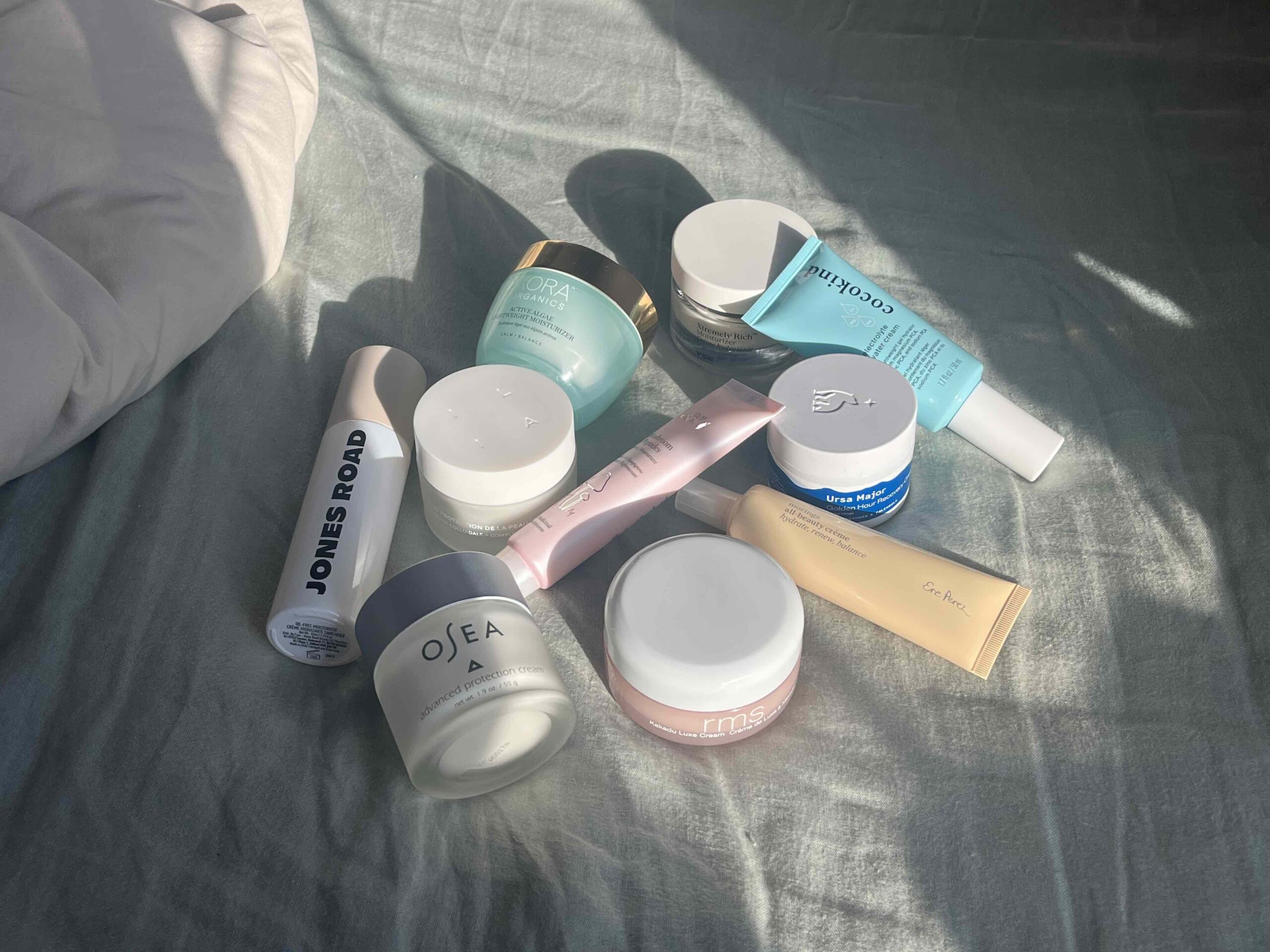 A collection of skincare and beauty products, including jars and tubes, arranged on a bed with sunlight casting shadows.
