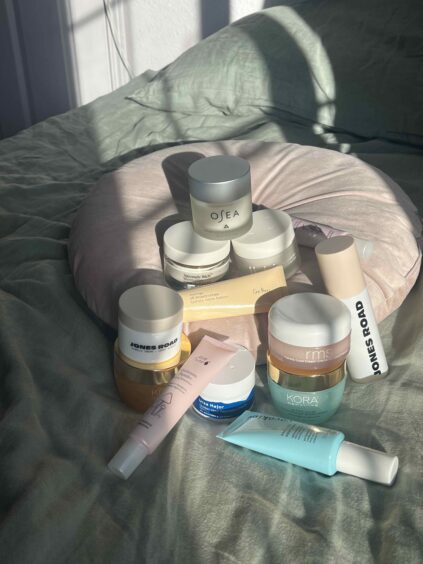 A collection of skincare products, including jars and tubes from brands like OSEA, Jones Road, and Kora, arranged on a bed with a round pink pillow in sunlight.