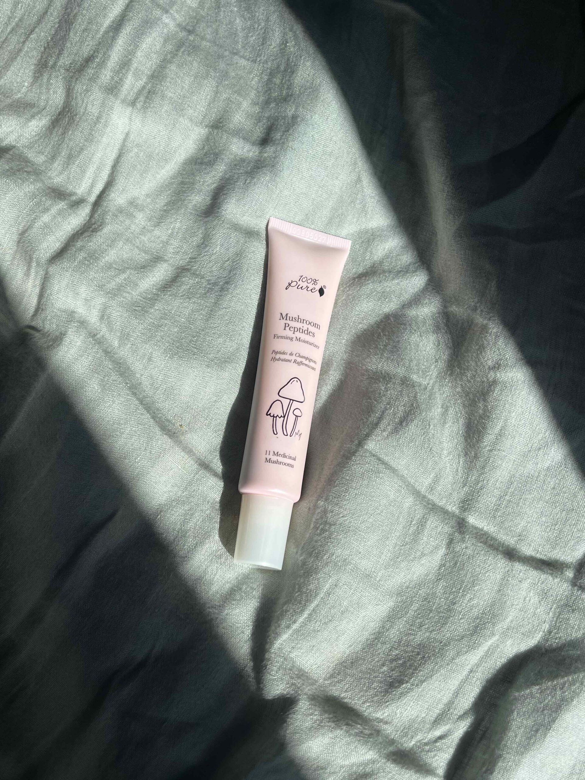 A tube of Fiddy&Friends Mushroom Peptide cream lies on a light green, crumpled fabric, with sunlight casting shadows across the scene.