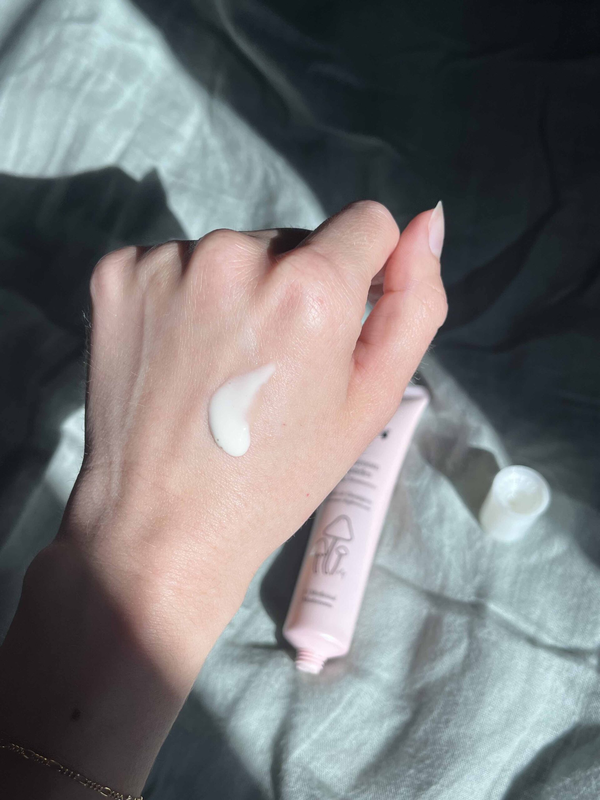 A hand with a dab of white cream on the back, holding a pink tube of skincare product with the cap off, on a light fabric background.