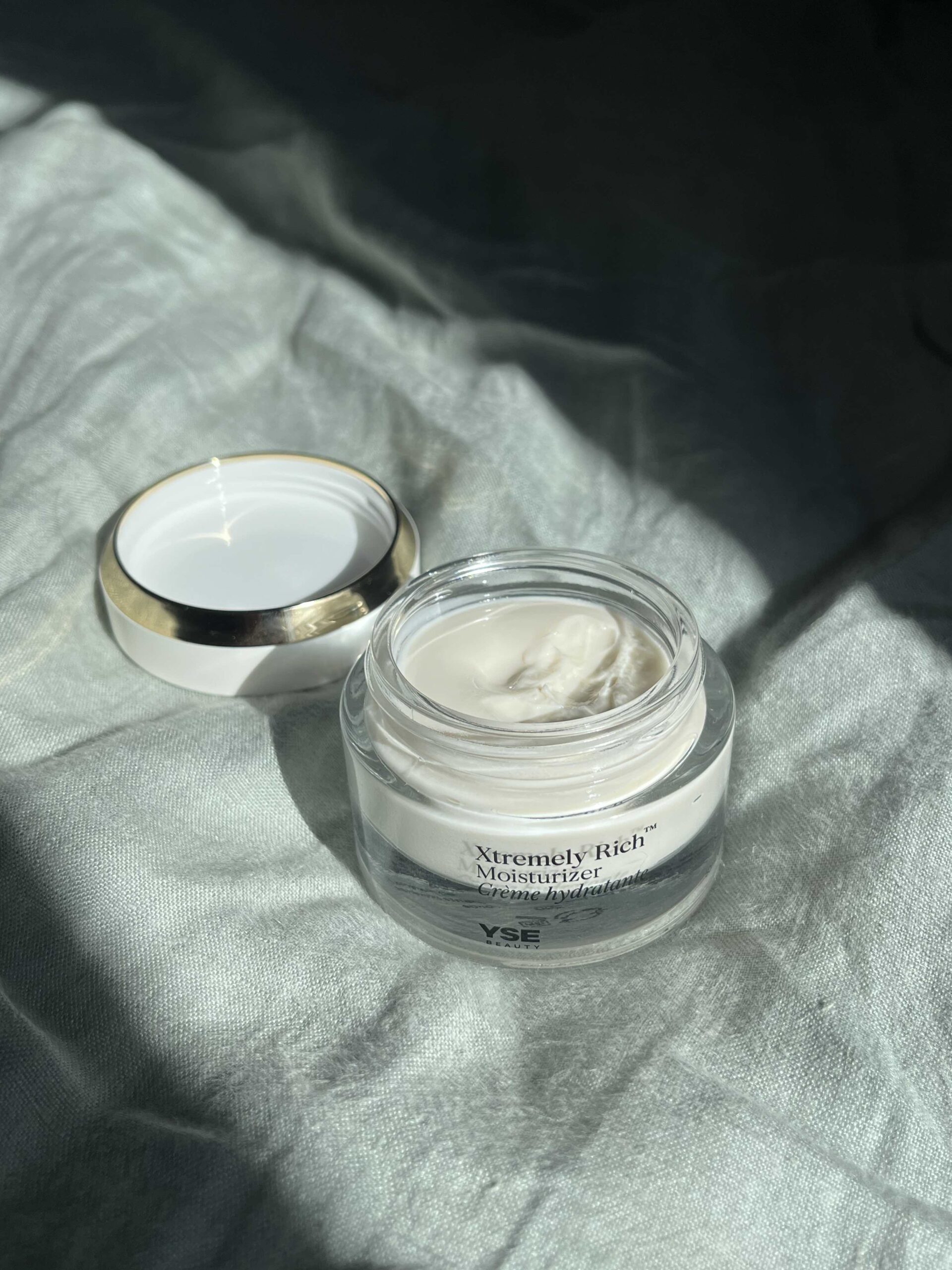 An open jar of YSE Xtremely Rich Moisturizer with the lid placed beside it, sitting on a textured light fabric surface under soft lighting.