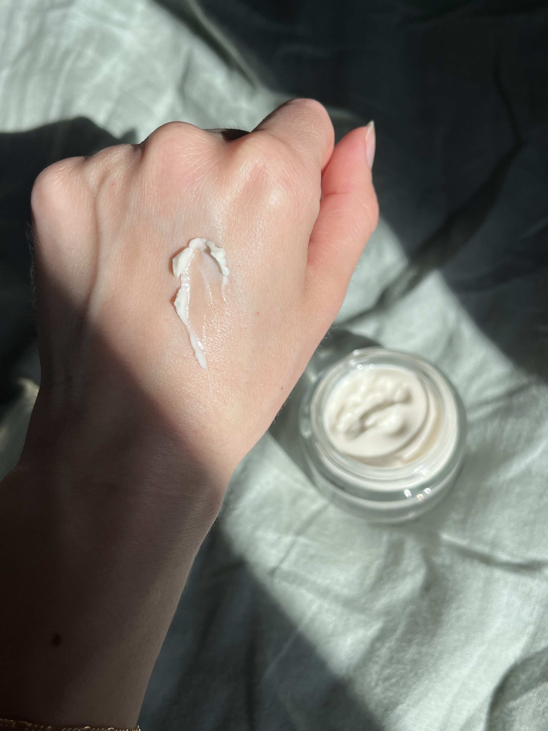 A hand with a dab of white cream on the back is held above an open jar of cream, both on a light fabric background.