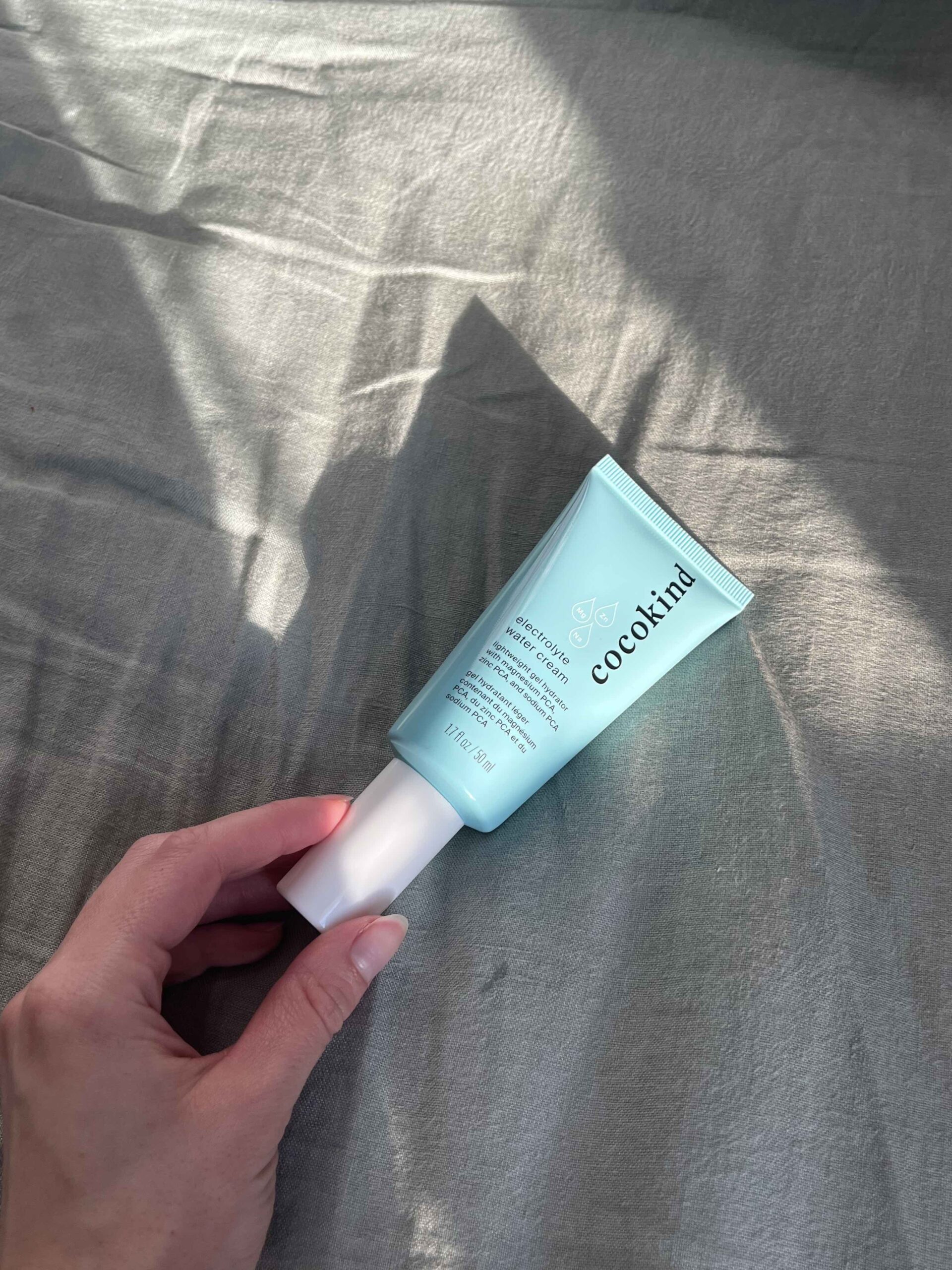 A hand holding a tube of Cocokind skin care product on a light gray fabric surface with soft natural lighting.