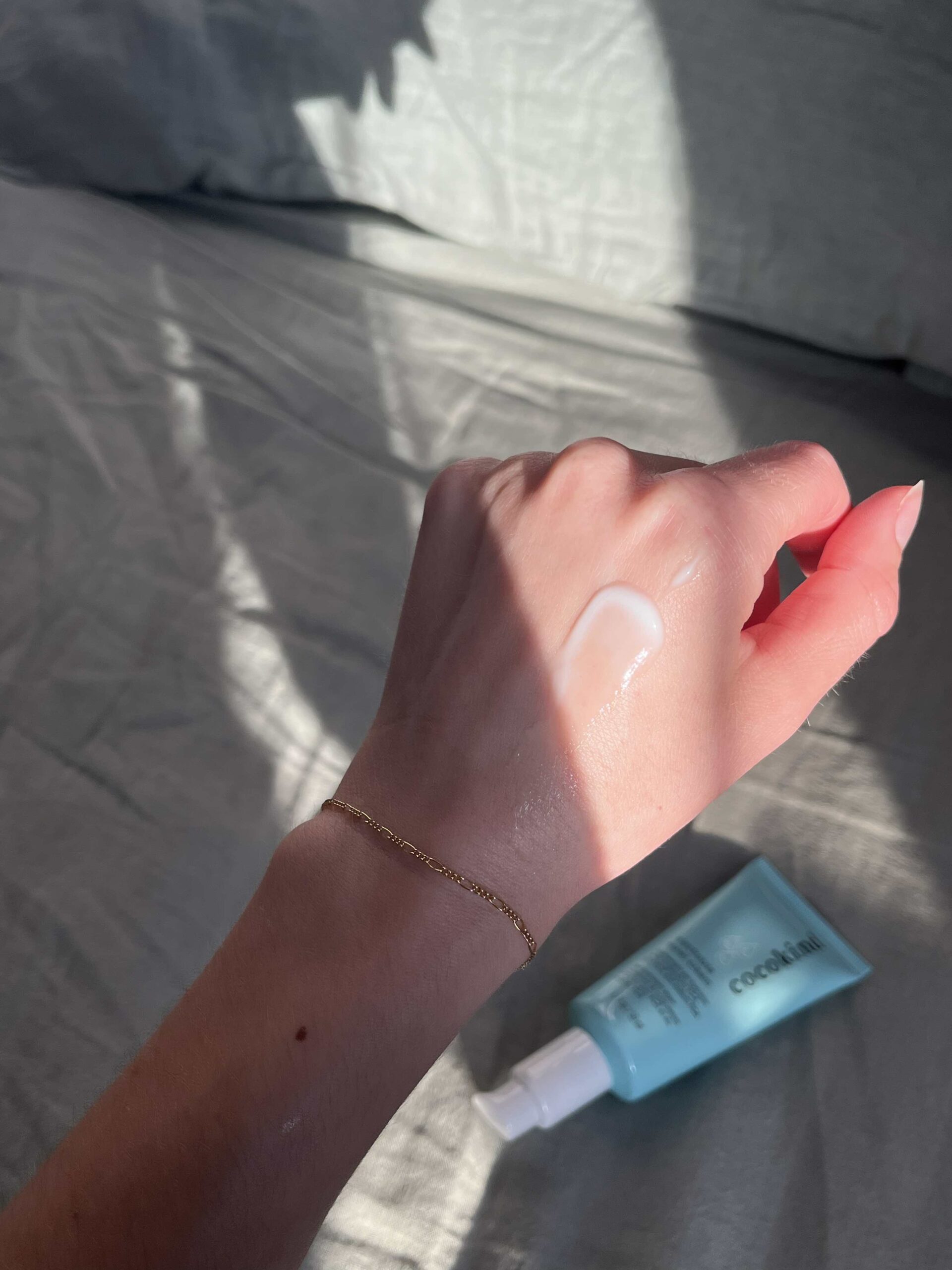 A hand with a gold bracelet has a dab of white cream on the back; a blue tube labeled "coola" is nearby on a light surface.