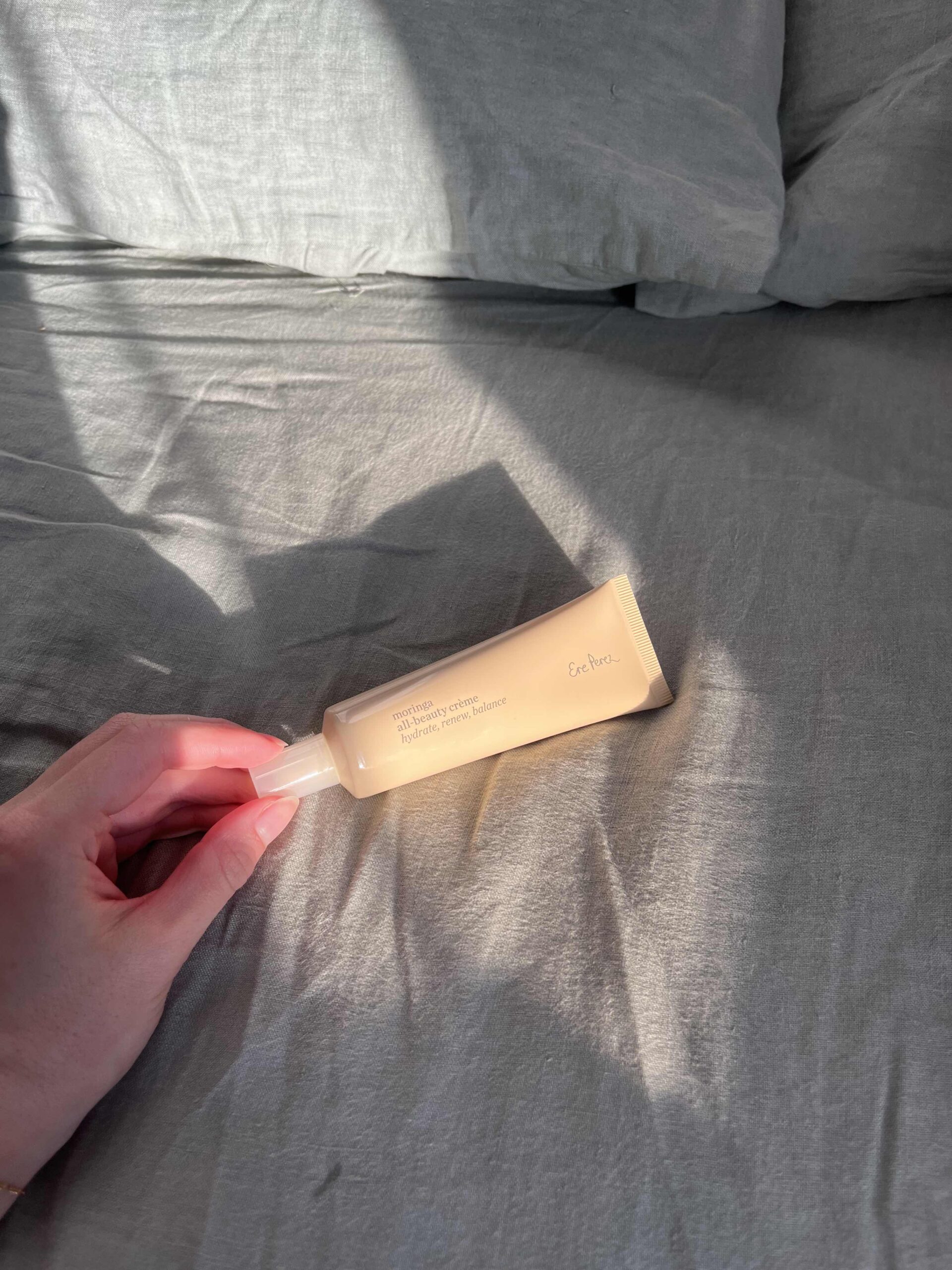 A hand holding a beige tube of skincare product labeled "Everyday Sunscreen" on a gray bedsheet with sunlight casting shadows.