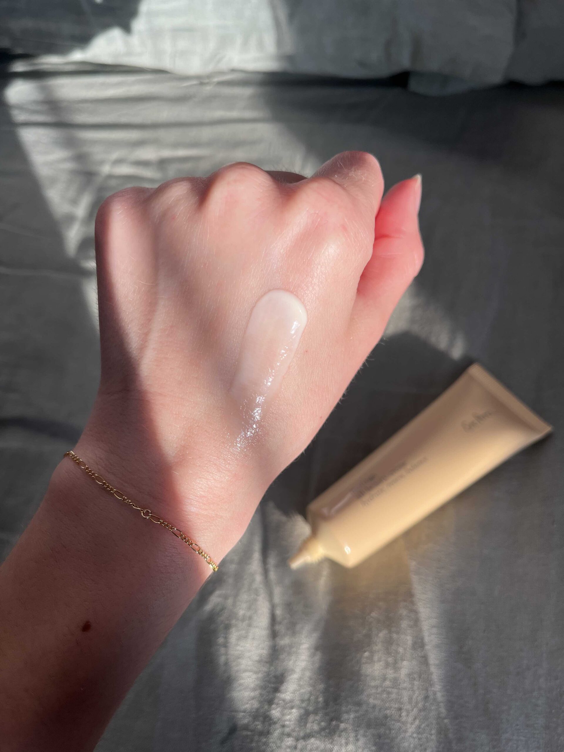 A hand with a gold bracelet shows a swipe of translucent cream on the back, with a beige tube of cream lying nearby on a gray surface.
