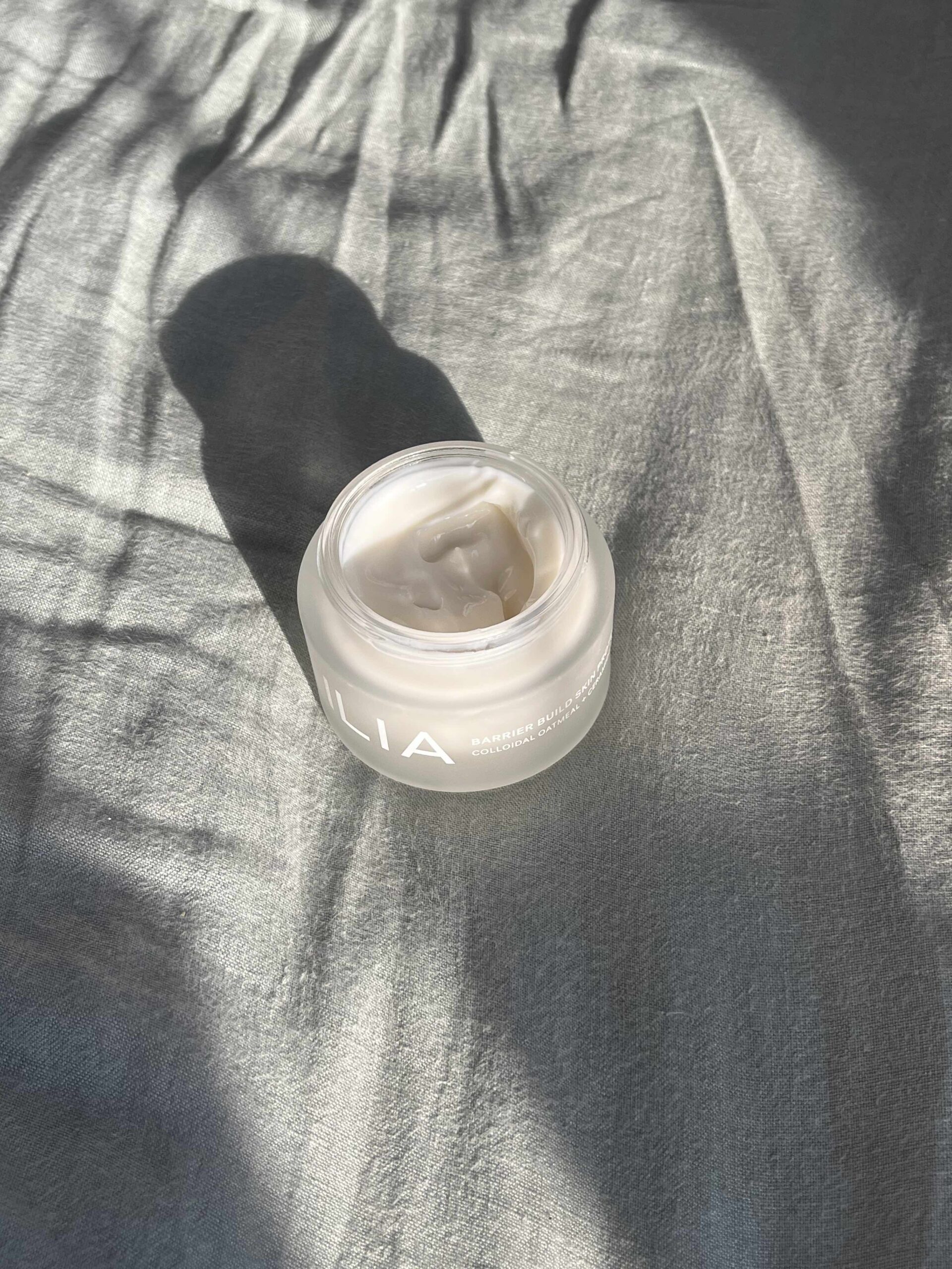 A glass jar of white cream sits on a textured, gray fabric surface in natural sunlight, casting a soft shadow.
