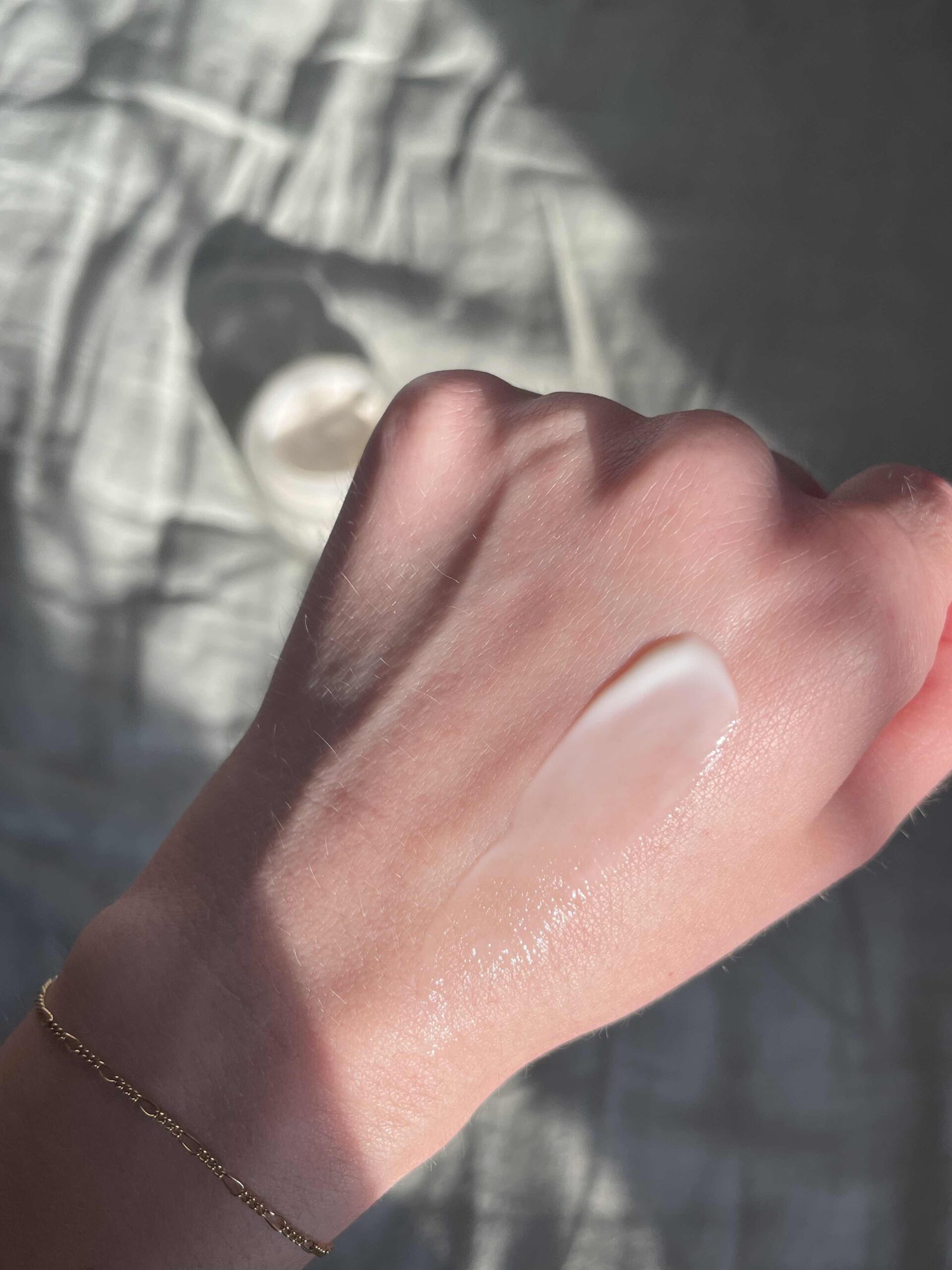 A hand with a gold bracelet shows a swipe of white cream or lotion applied to the back of it, with a jar of cream blurred in the background.