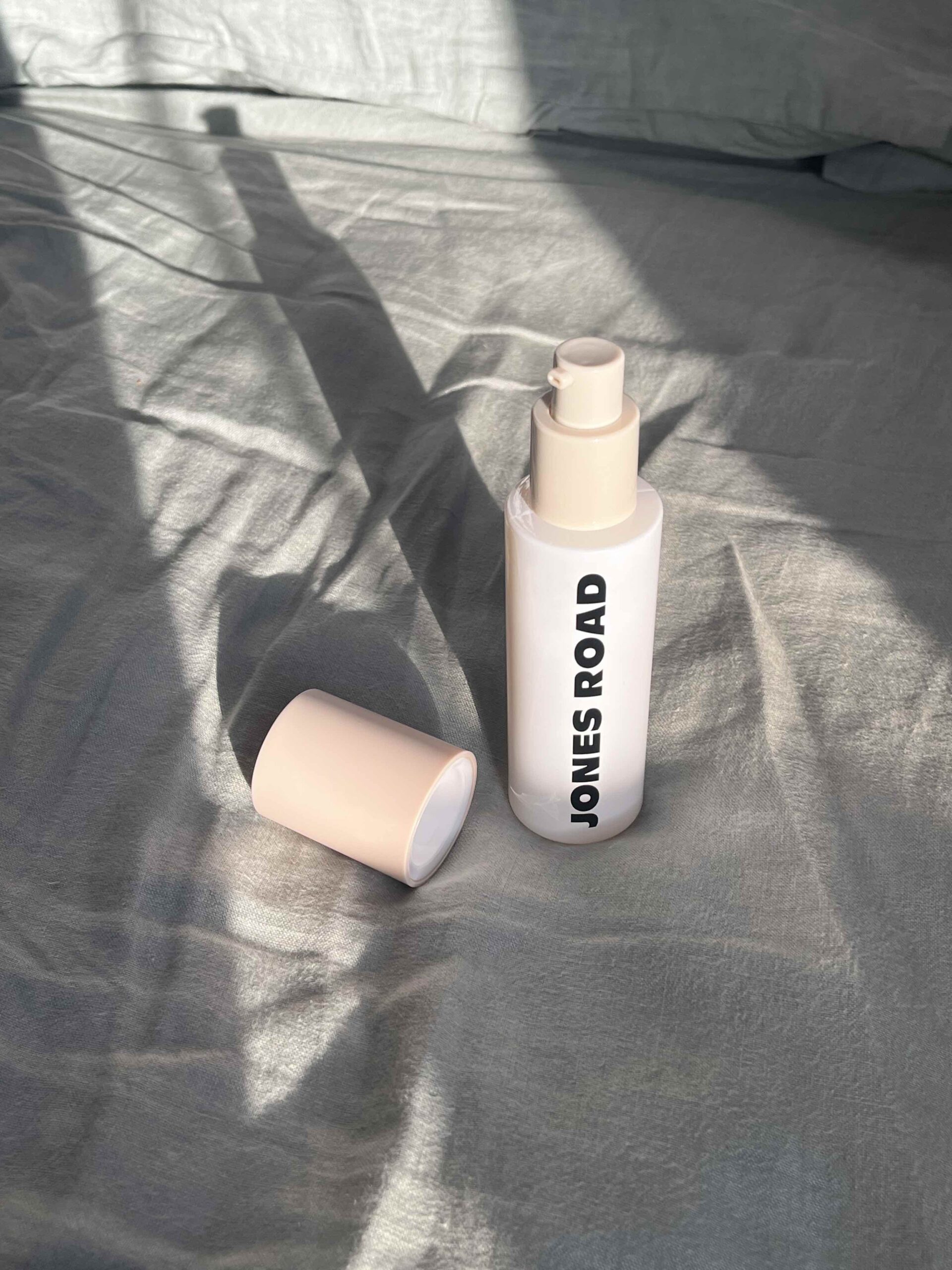 A bottle labeled "Jones Road" with a pump dispenser and a detached cap is placed on a textured gray fabric surface.
