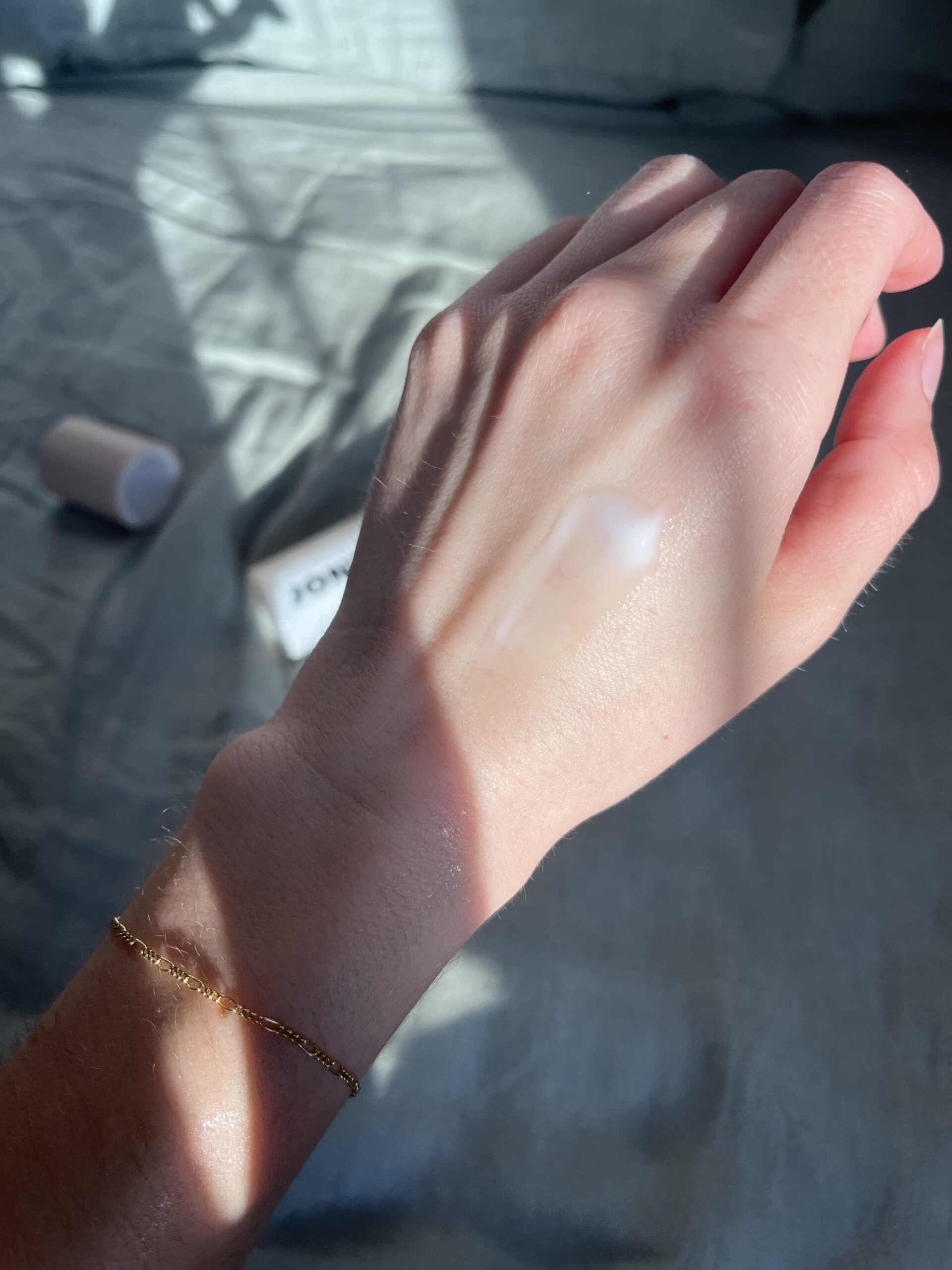 A close-up of a hand with a streak of white cream on the back, with sunlight casting shadows and a small gold bracelet on the wrist. Blurred objects are in the background.