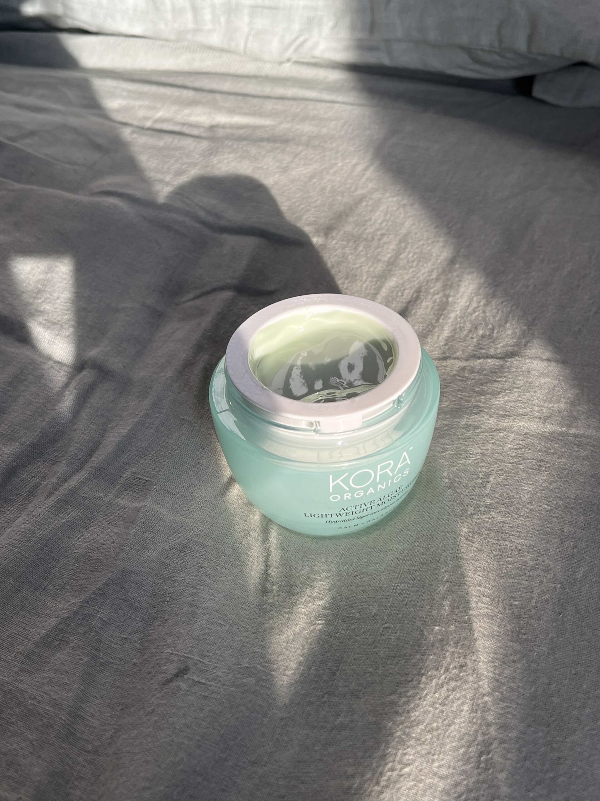 A jar of Kora Organics Turmeric Glow Moisturizer with the lid off sits on a gray fabric surface in natural sunlight.