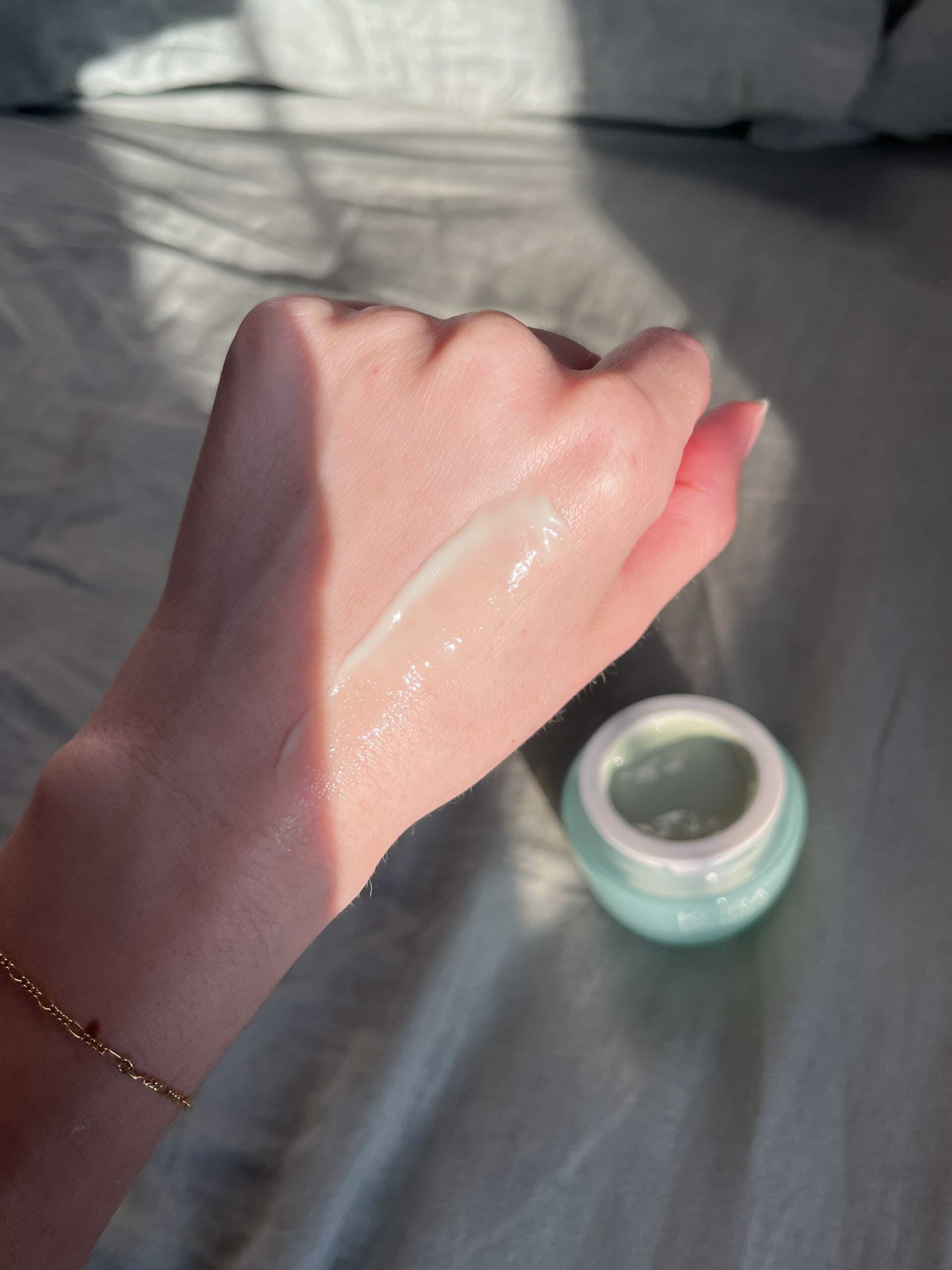 A hand with a swipe of cream on the back is shown next to an open jar of light green cream, with sunlight casting shadows across the scene.