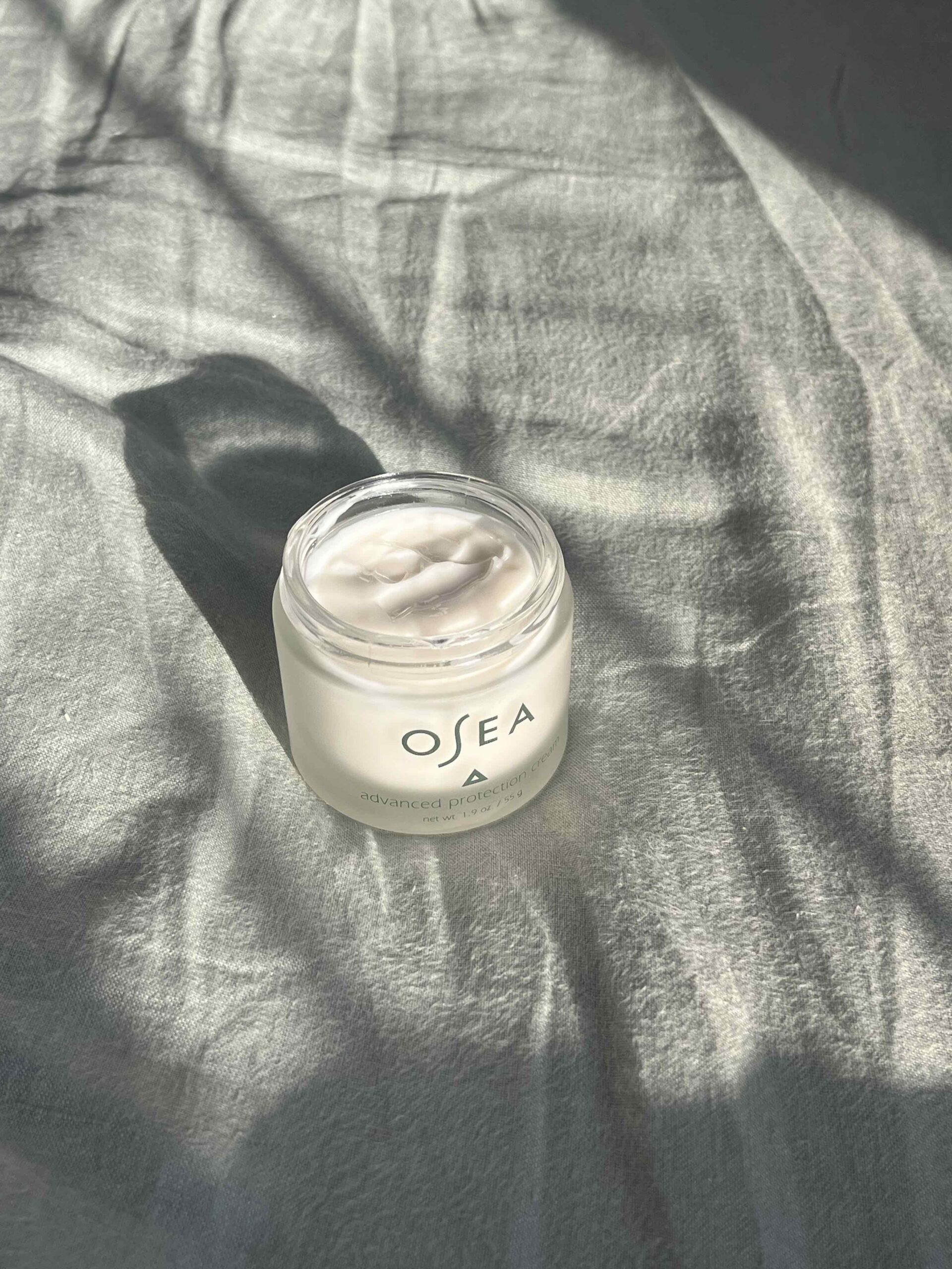 A jar of OSEA Advanced Protection Cream sits on a wrinkled, sunlit fabric surface, casting distinct shadows.