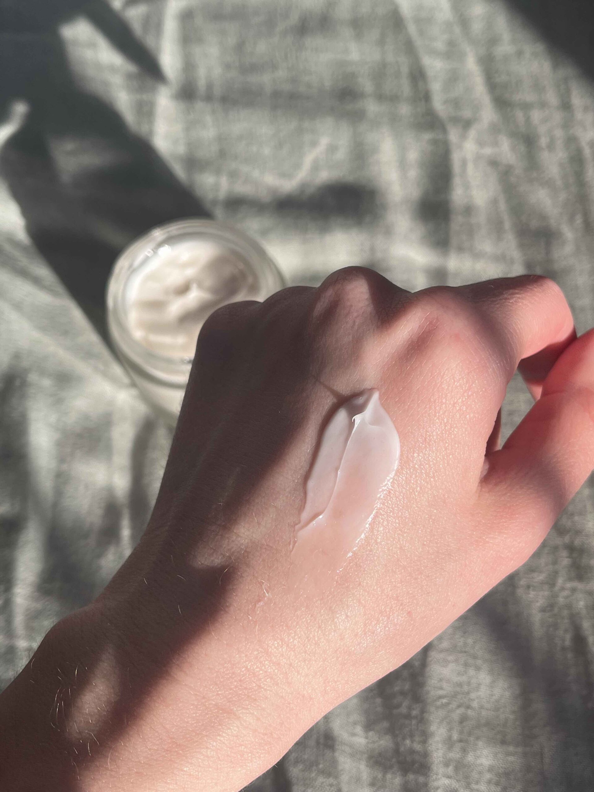 A hand with a streak of white cream on the back, positioned above a jar of cream on a fabric surface in natural light.