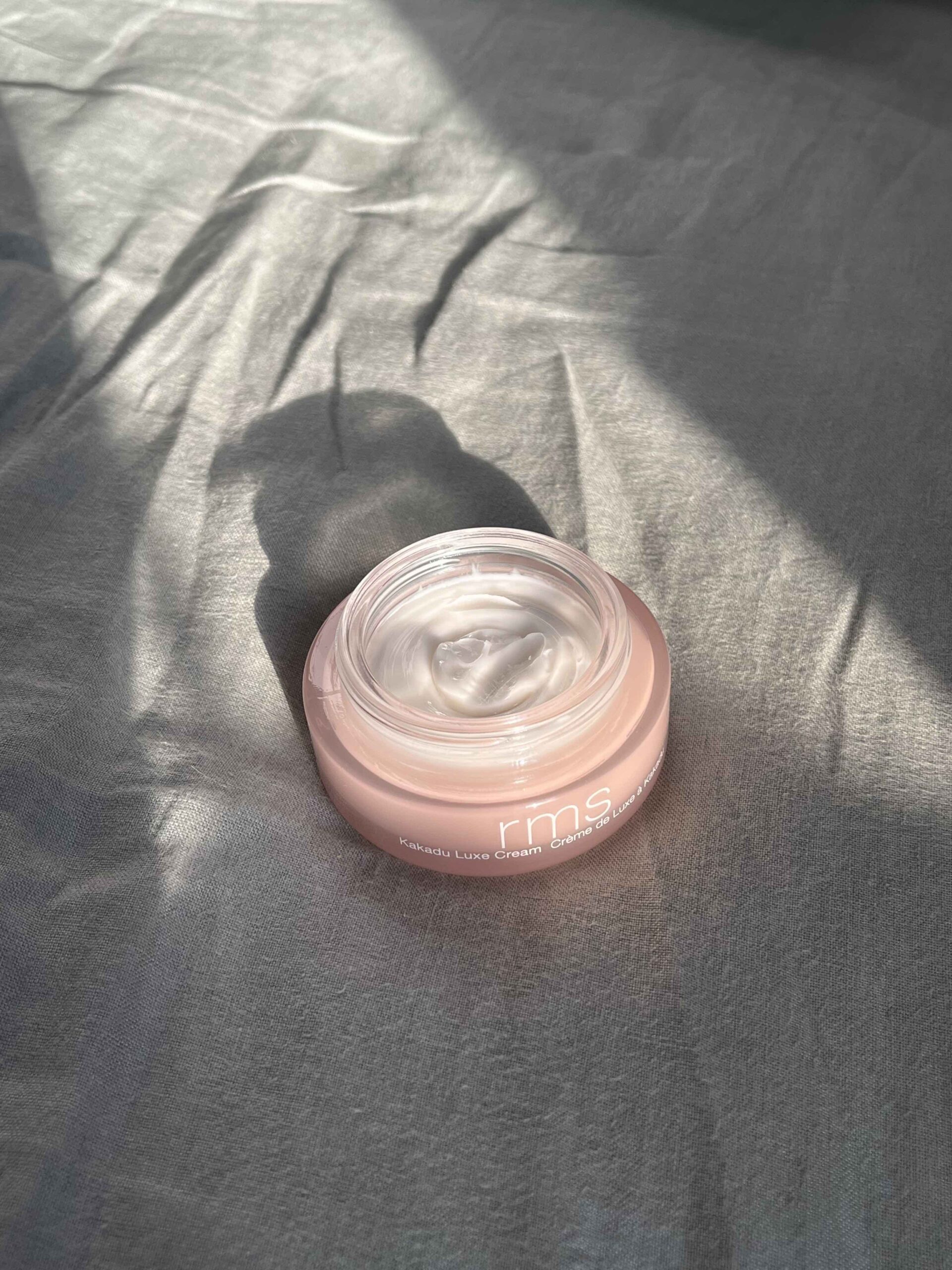 An open jar of RMS Kakadu Luxe Cream sits on a gray fabric surface, illuminated by sunlight streaming from the side.