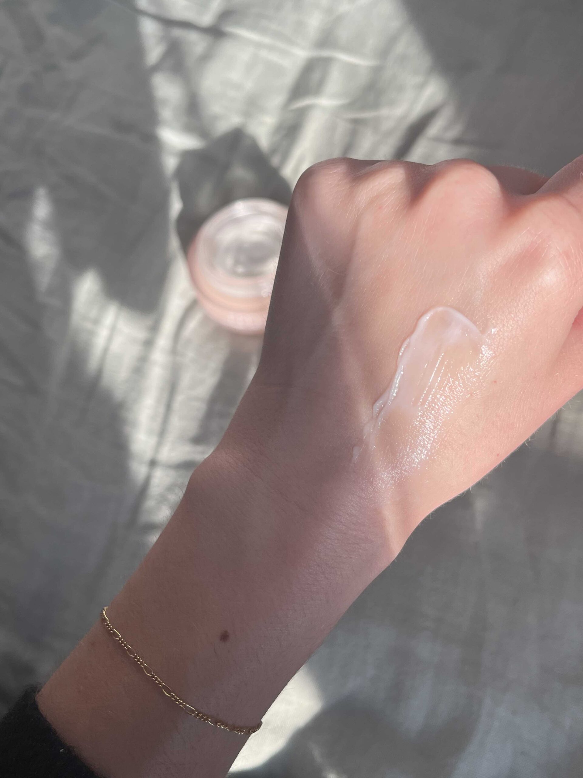 A hand with a gold bracelet has a streak of white cream applied to the back, with a jar of cream visible in the background on a fabric surface.
