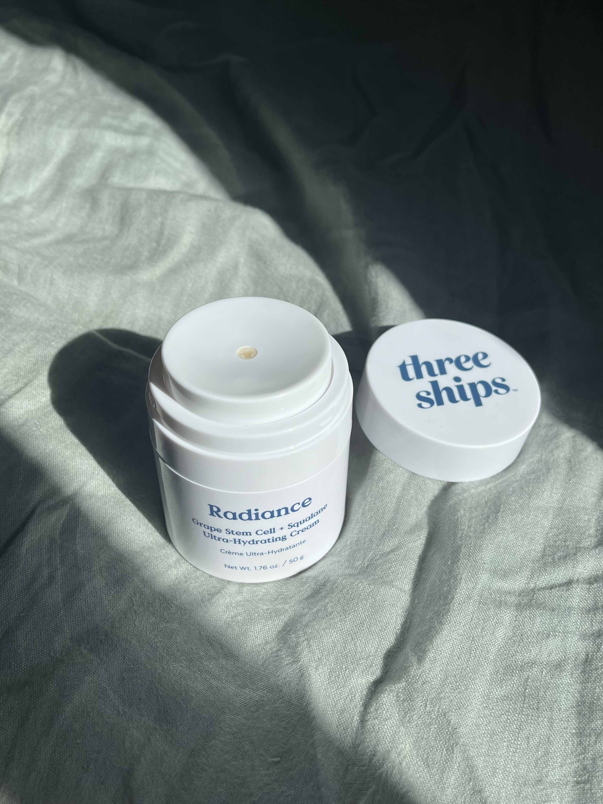 A white container of "Radiance Grape Stem Cell + Squalane Day Cream" by Three Ships sits on a fabric surface with its lid placed beside it.