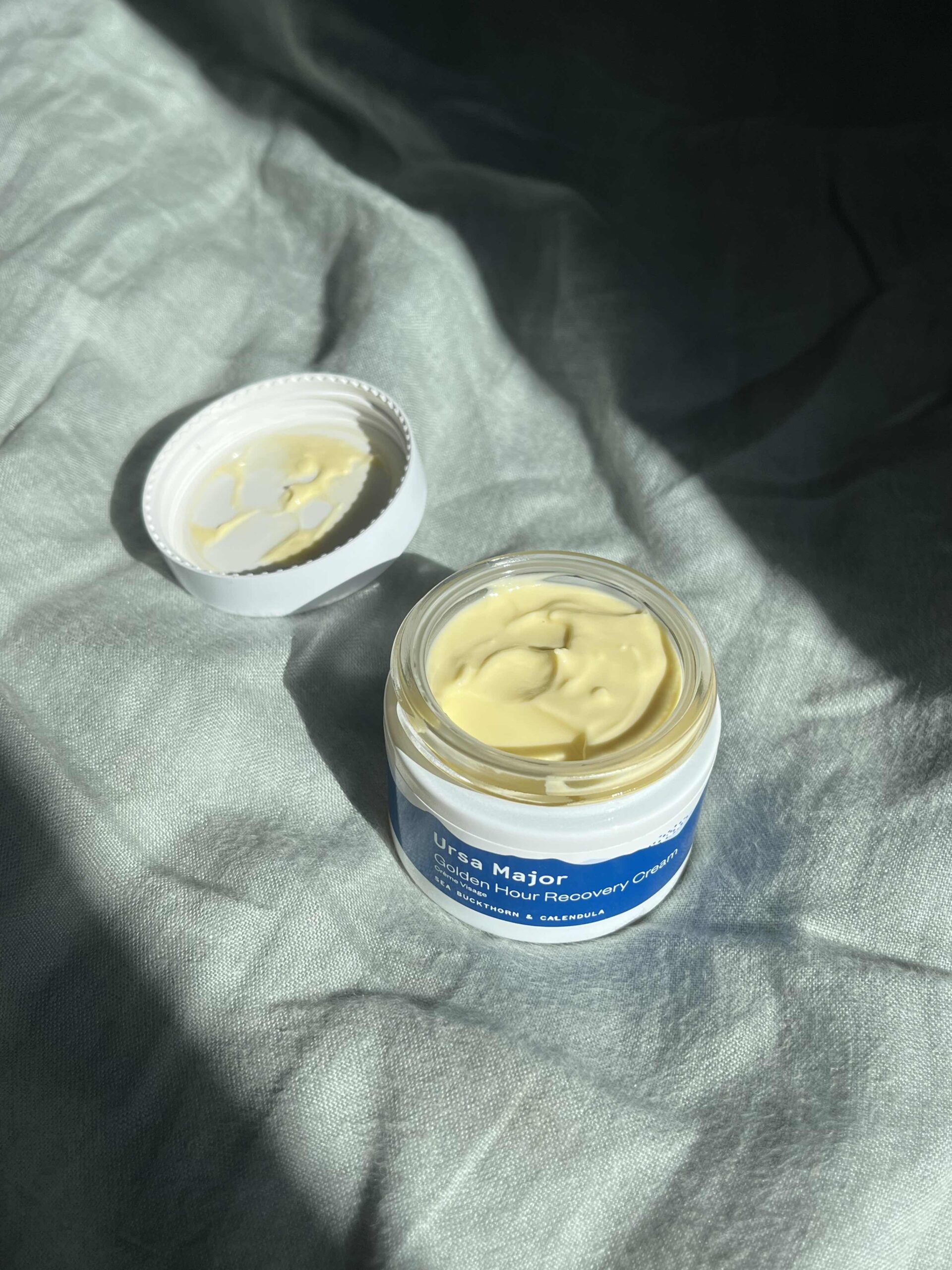 An open jar of Ursa Major Golden Hour Recovery Cream sits on a textured fabric surface, with the white lid placed beside it.