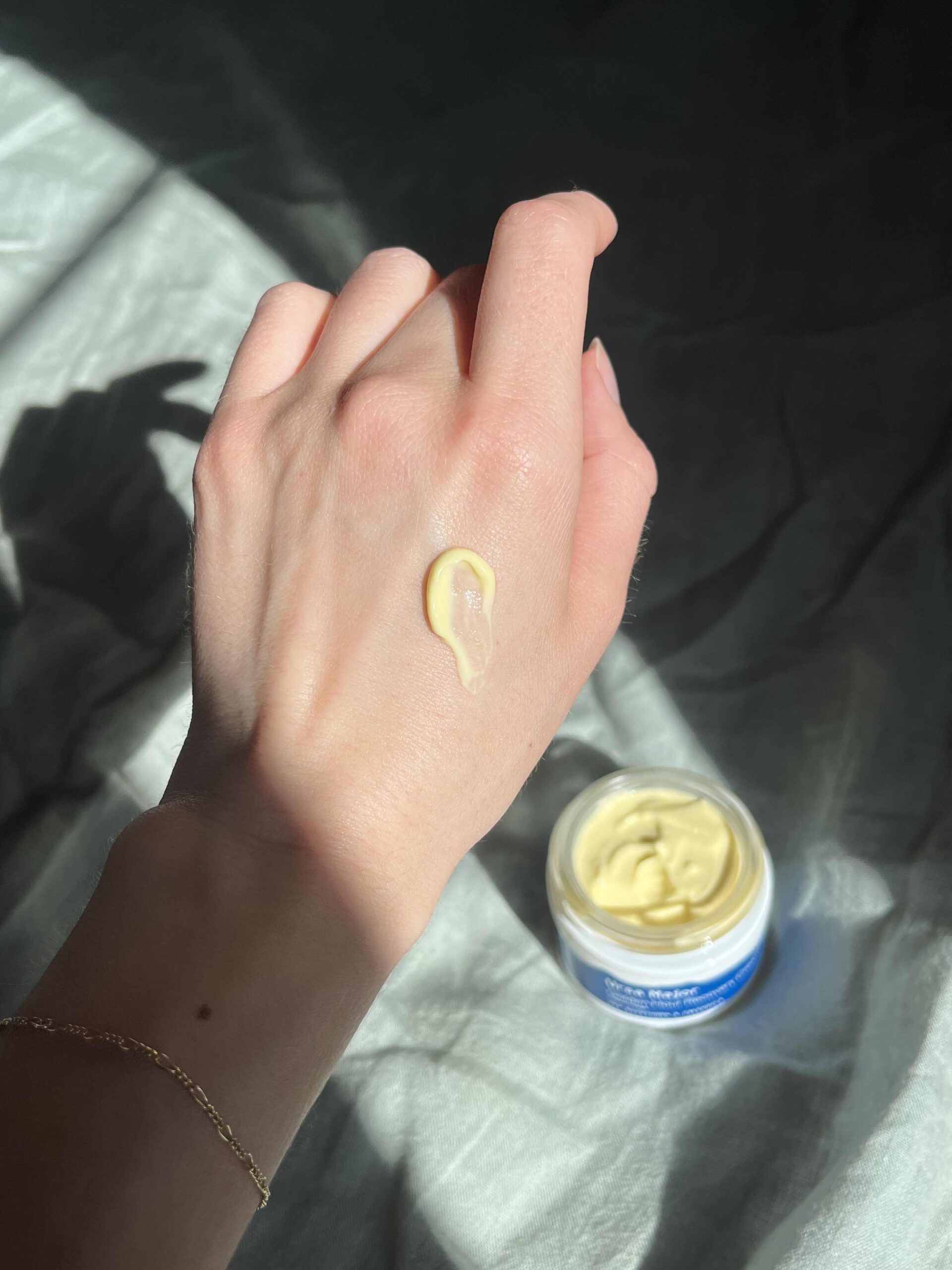 A hand with a gold bracelet shows a smear of yellow cream on the back, next to an open jar of cream on a light fabric surface.