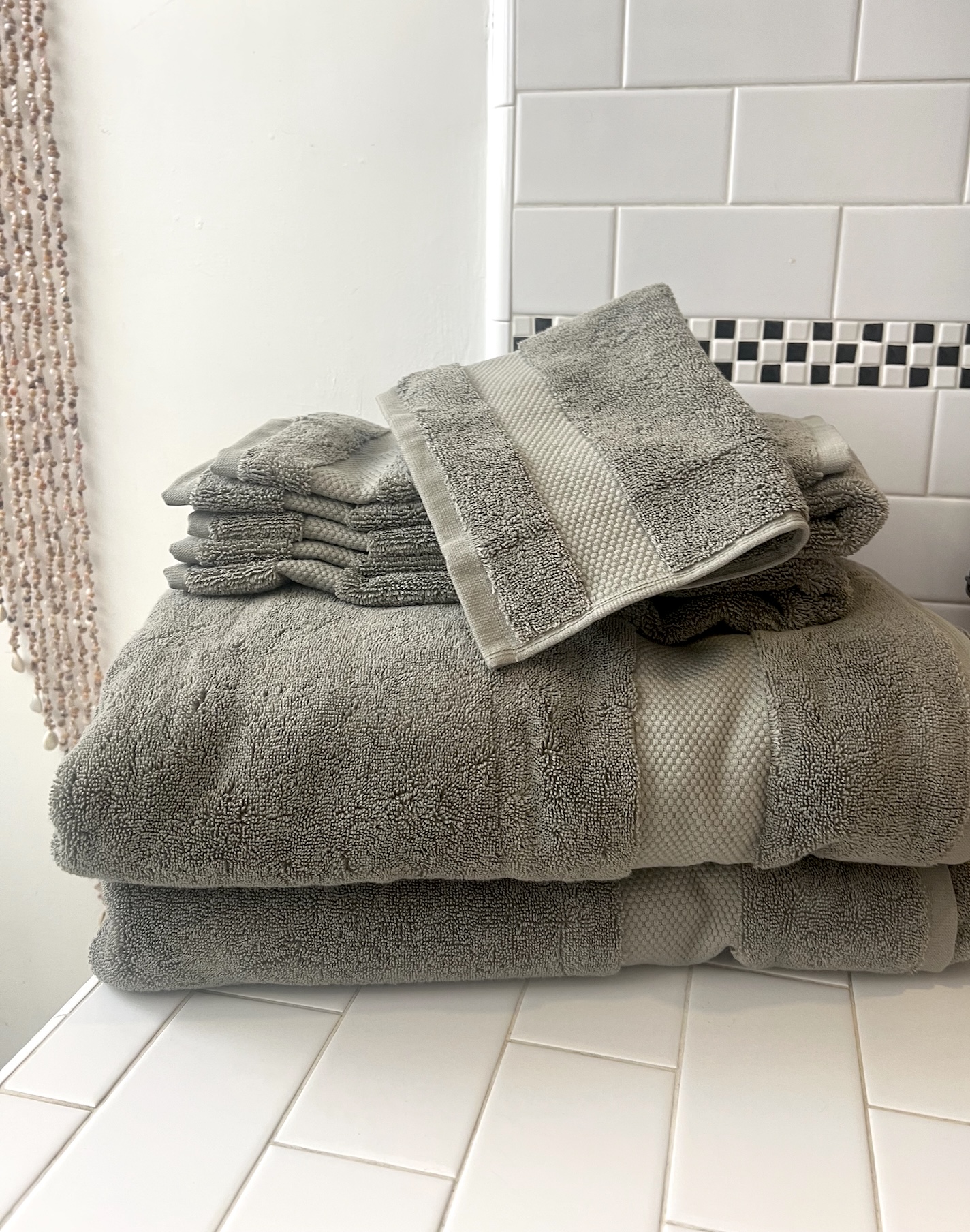 A neatly folded stack of organic towels in various sizes sits on a white tiled bathroom countertop near a beaded curtain and white tiled wall.