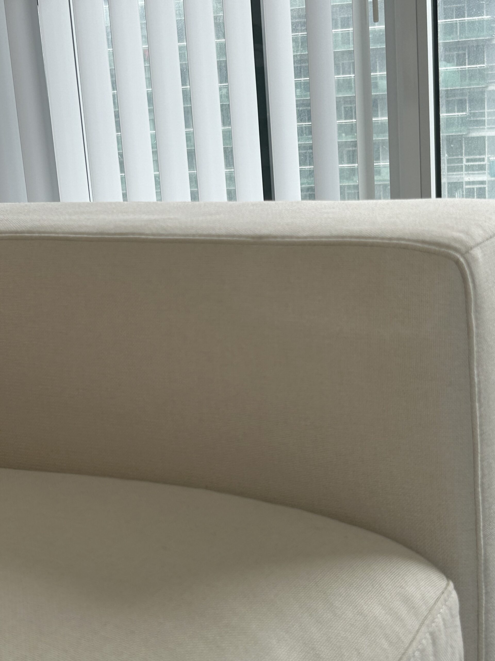 A close-up of a beige upholstered couch in front of vertical white blinds, with a window and city buildings visible in the background.