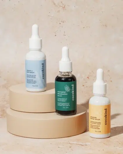 Three dropper bottles of serum, each with different colored labels, are displayed on stacked beige circular platforms against a neutral background.