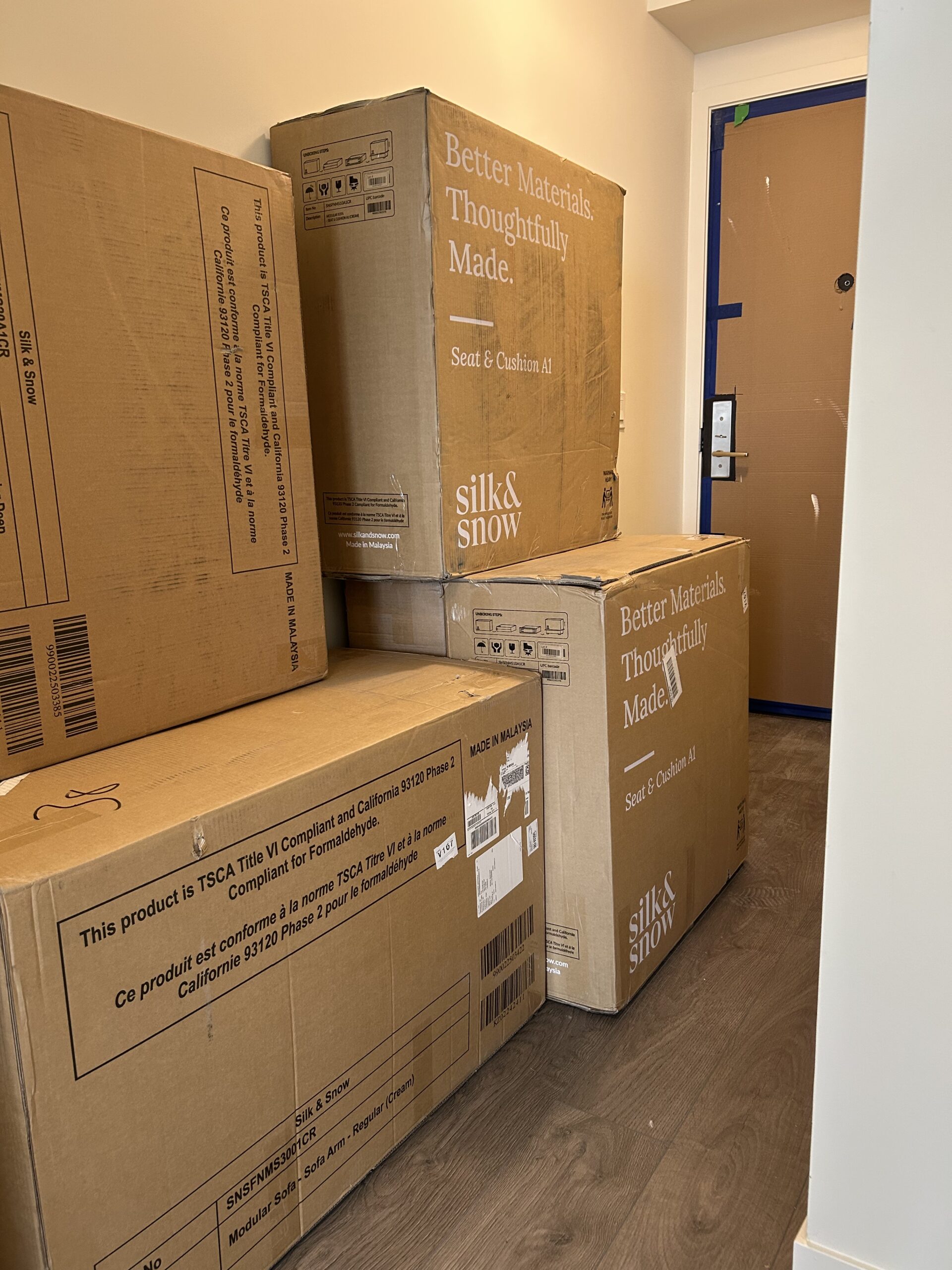 Several large cardboard boxes labeled "silk & snow" stacked near a doorway in a room with wooden flooring and a partially covered door.