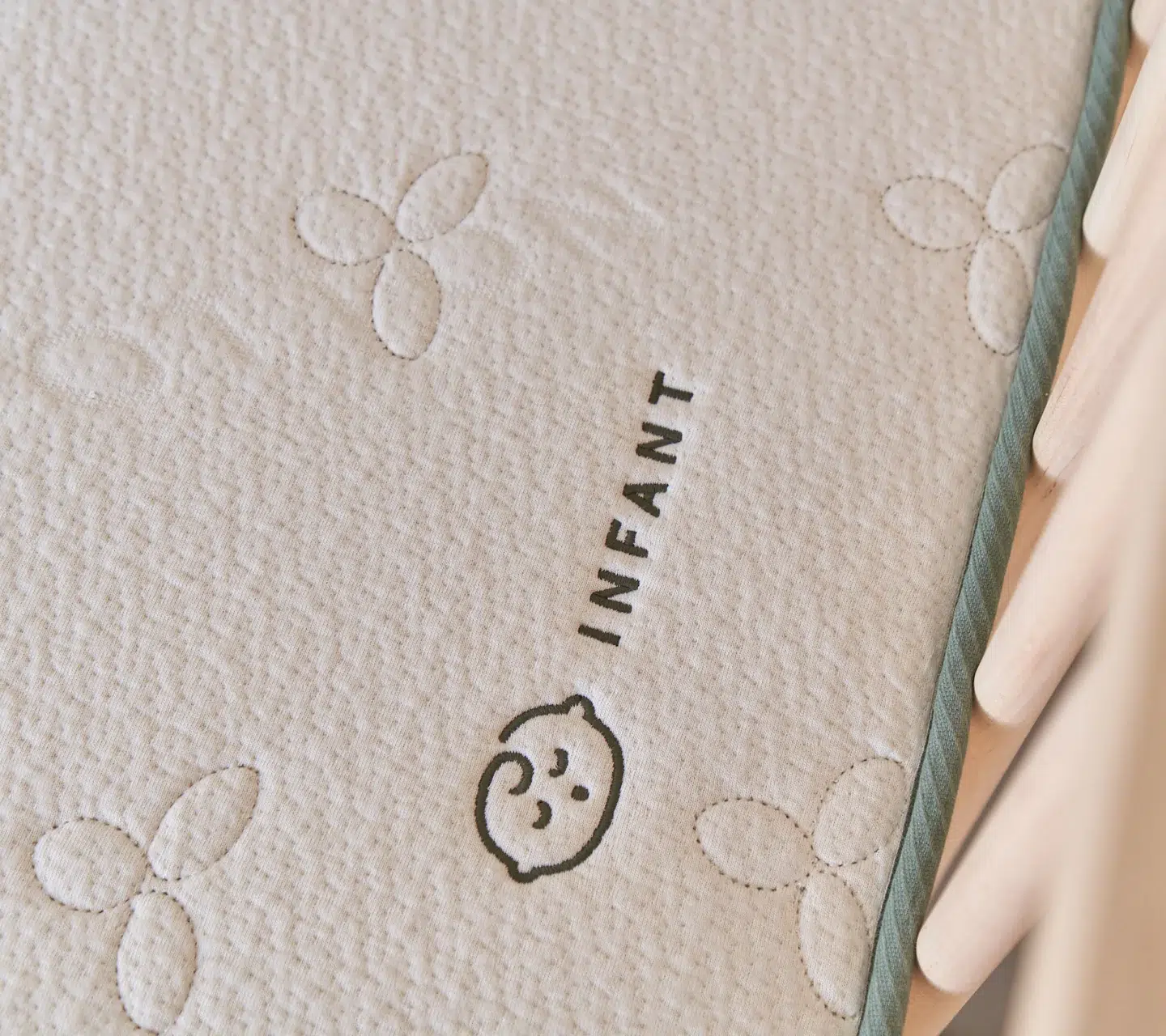 Close-up of a mattress labeled "INFANT" with an outline of a baby’s face and quilted floral patterns, next to the wooden bars of a crib.
