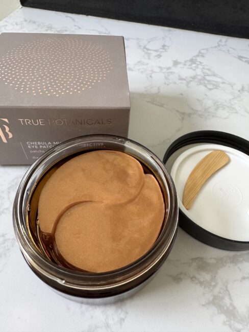 A jar of True Botanicals Chebula Melt Eye Masks with the lid off, showing gold-colored eye patches and a spatula on the open lid, next to the product box on a marble surface.