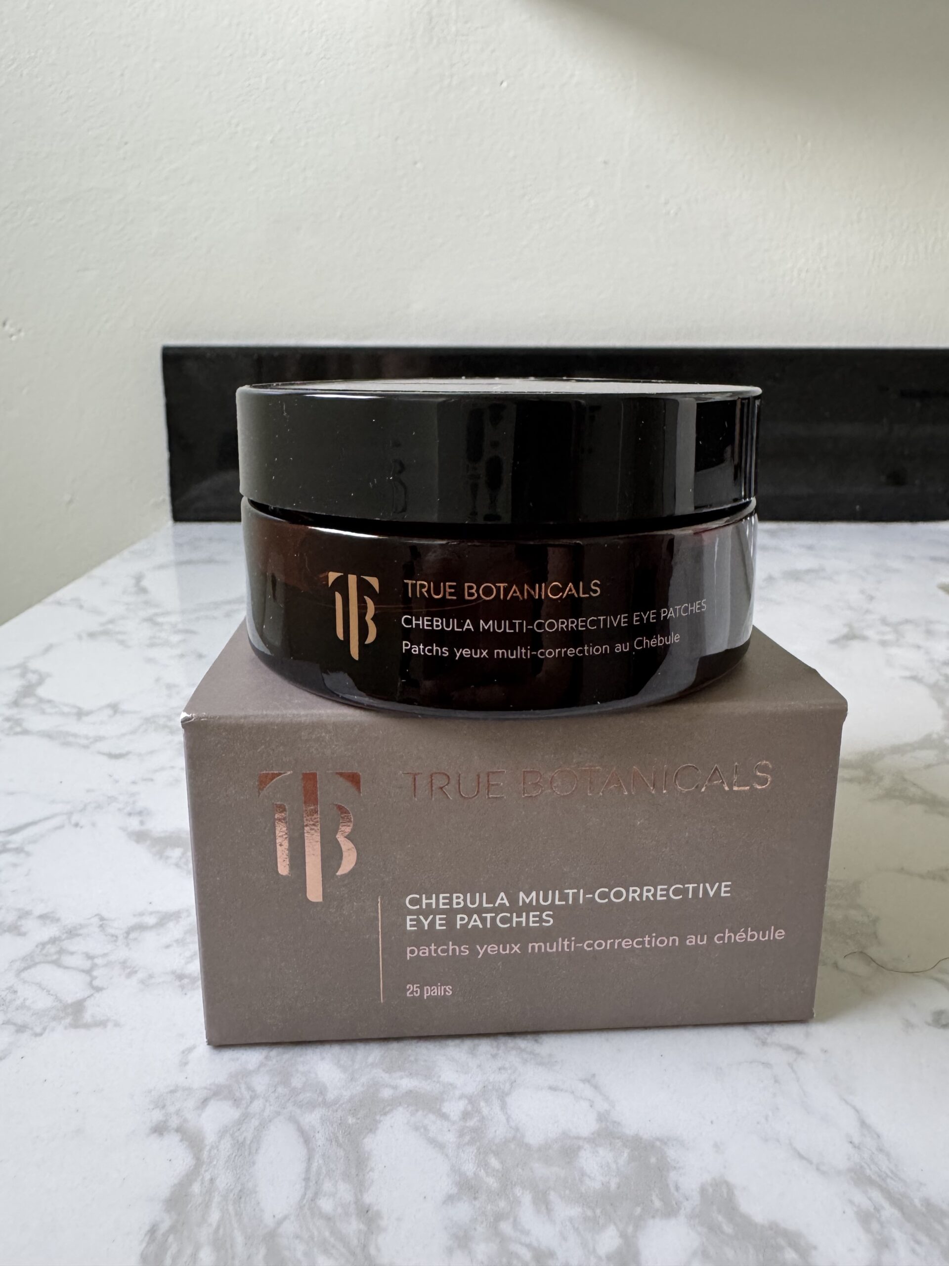 A jar of True Botanicals Chebula Multi-Corrective Eye Patches sits on top of its matching brown box on a white marble countertop.