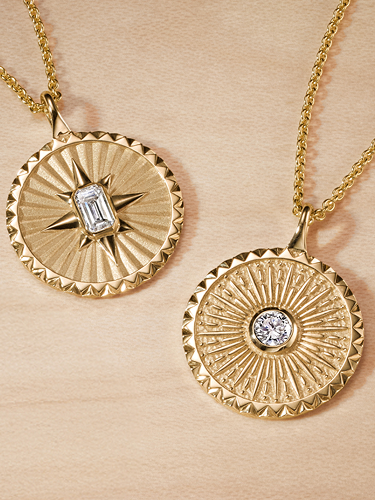 Two gold pendant necklaces on gold chains, each featuring a geometric sunburst pattern with a central clear gemstone, displayed on a light wood background.