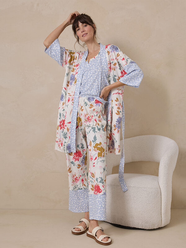 A woman stands indoors wearing a floral pajama set with a matching robe and sandals, posing beside a white textured chair against a beige wall.