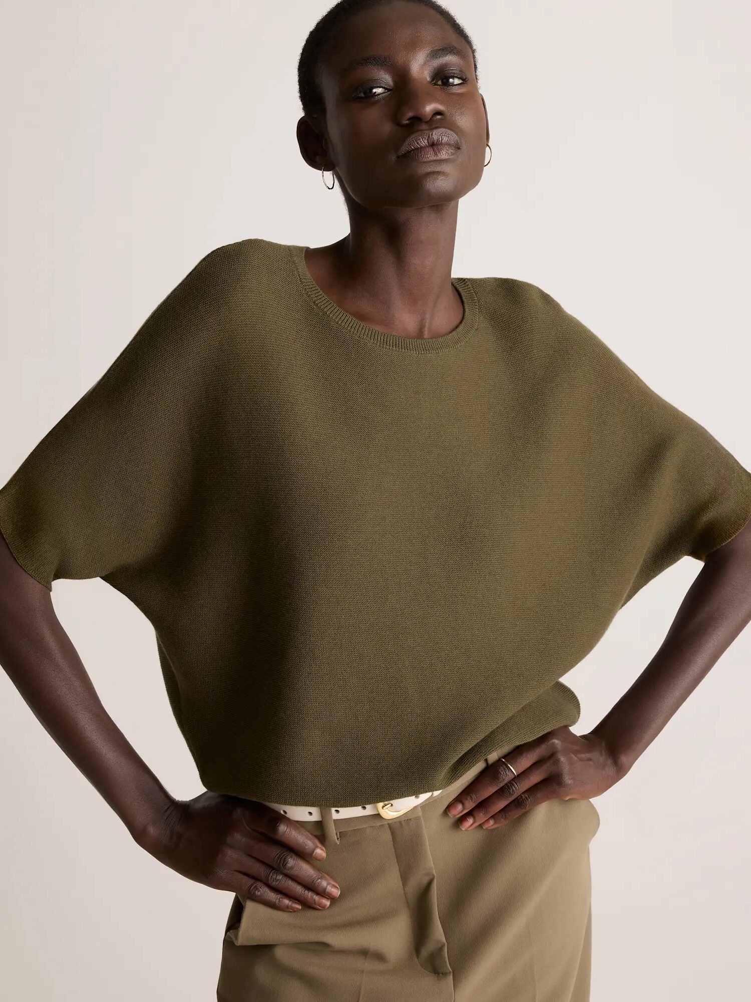 A person wearing an olive green, loose-fitting knit top and taupe pants stands with hands on hips against a plain background.
