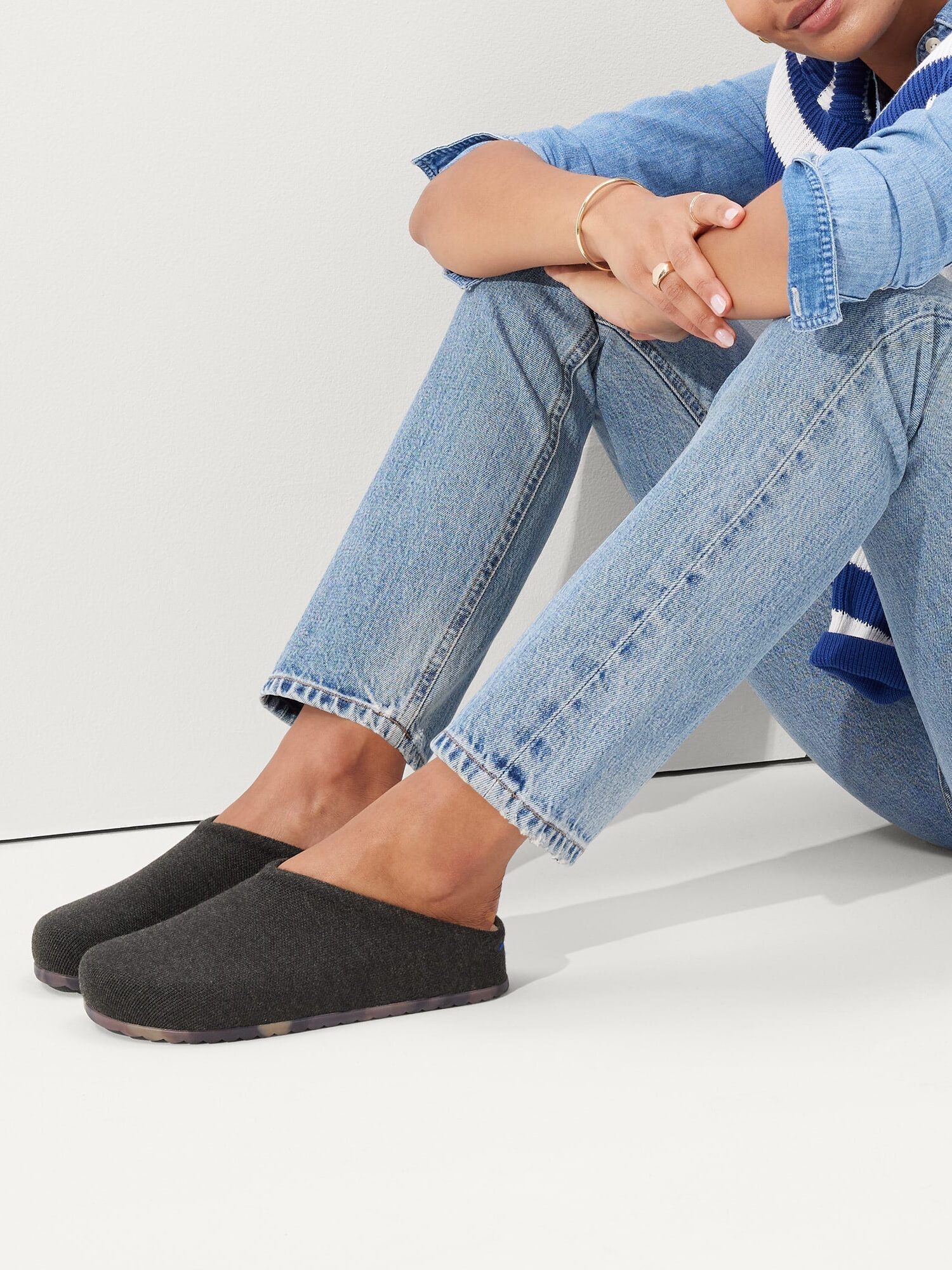 Person sitting on the floor with legs crossed, wearing light blue jeans, a denim shirt, a blue and white striped sweater, and dark slip-on clogs.