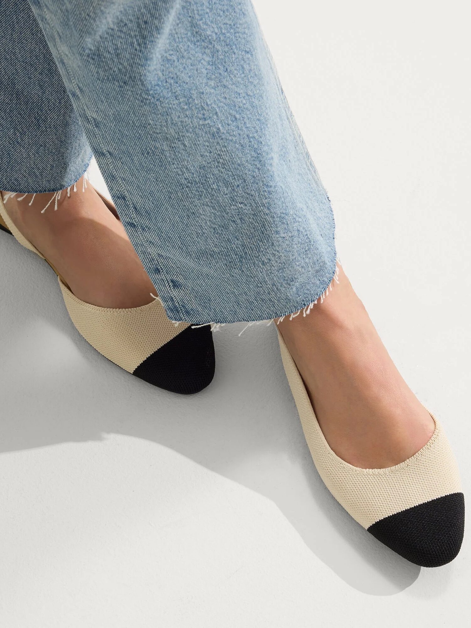 Person wearing light blue jeans and beige flats with black toe caps, standing on a light-colored floor.