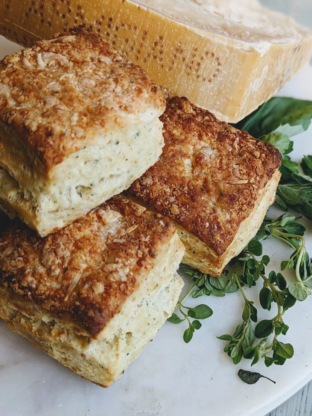 Three herb biscuits stacked on a white plate with fresh herbs and a wedge of hard cheese in the background.
