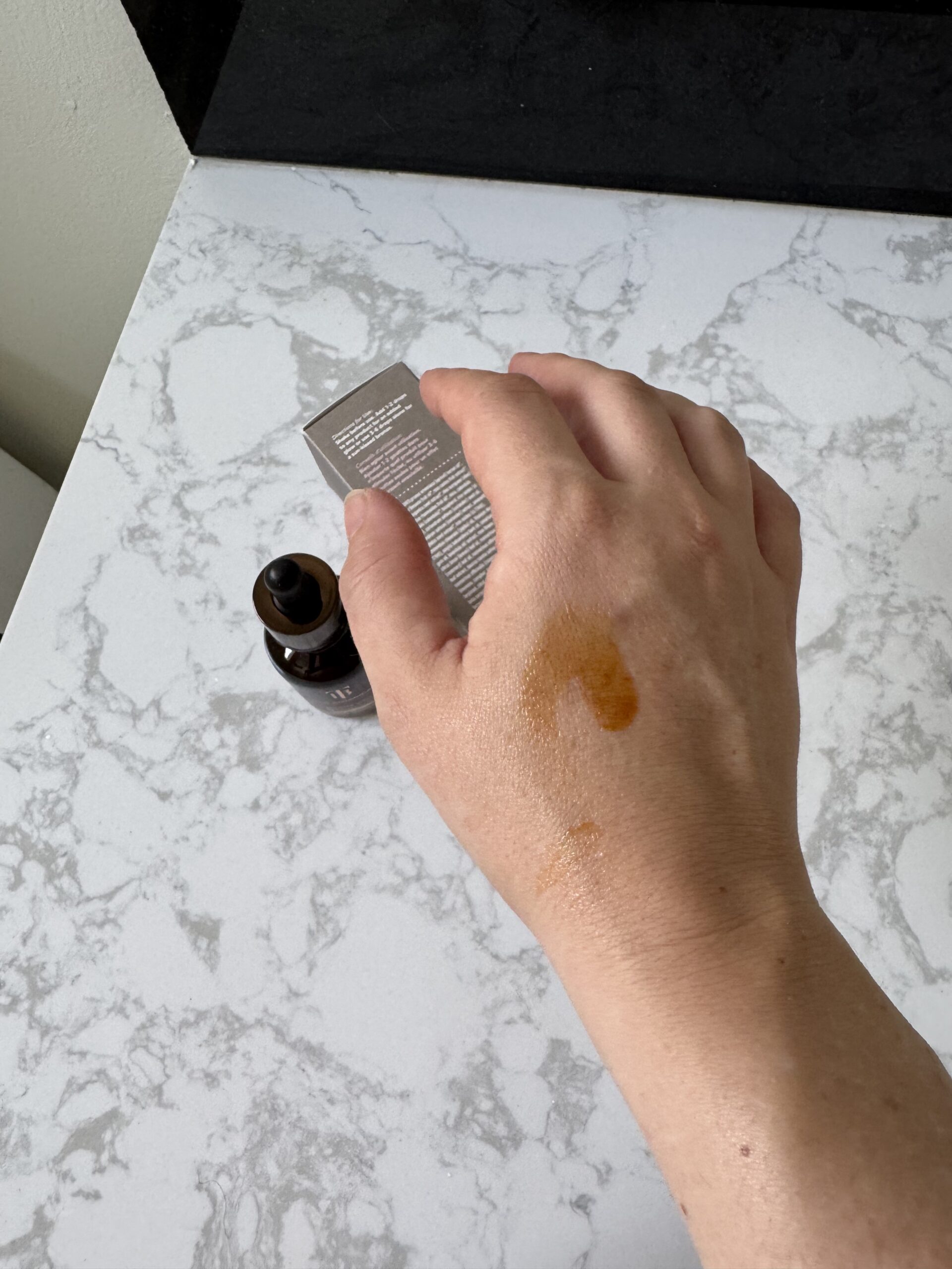 A hand with a patch of orange liquid on the back is shown above a marble surface, next to a dropper bottle and a small box.