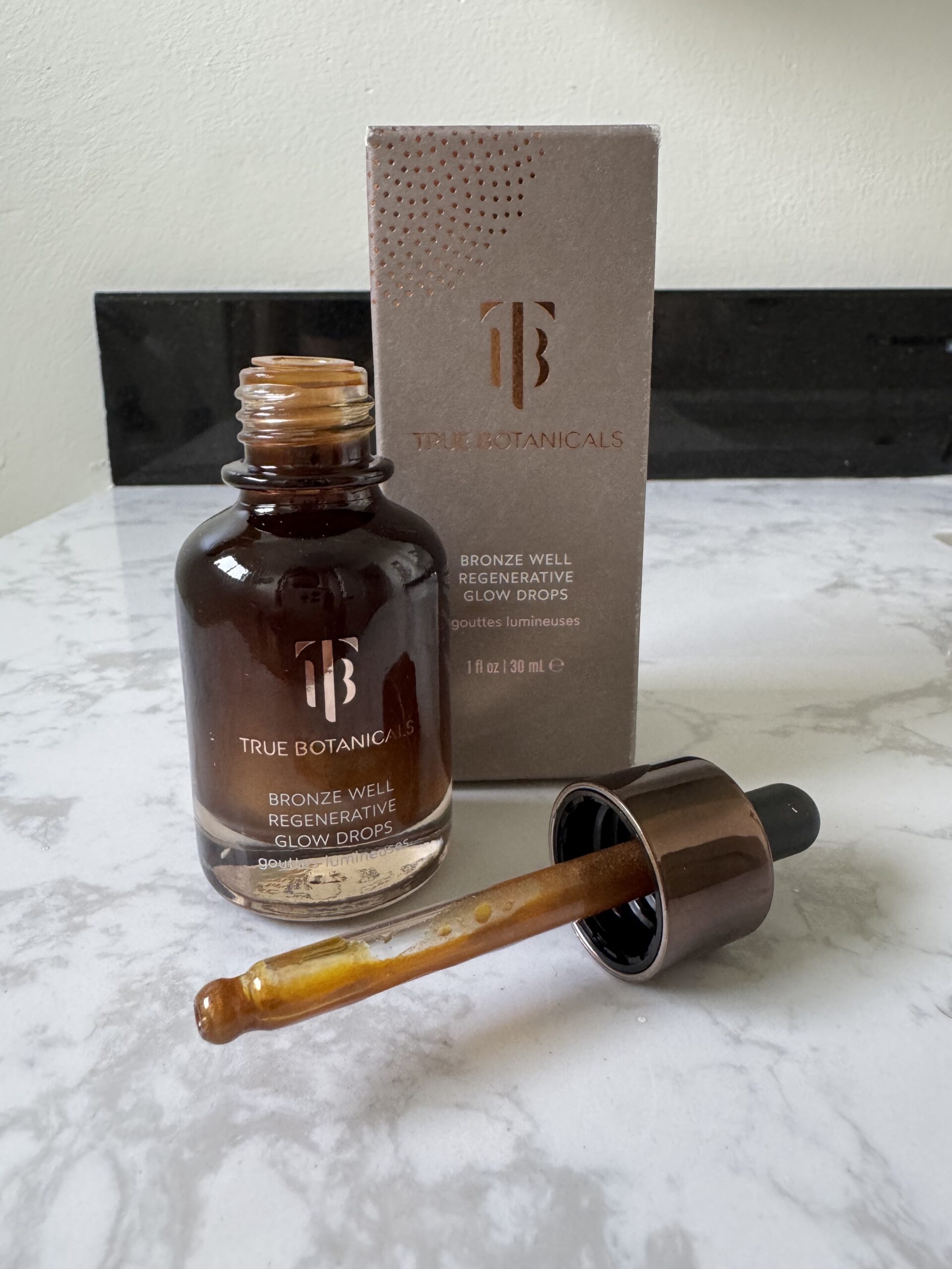A bottle of True Botanicals Bronze Well Regenerating Glow Drops with an open dropper, shown next to its packaging on a marble surface.