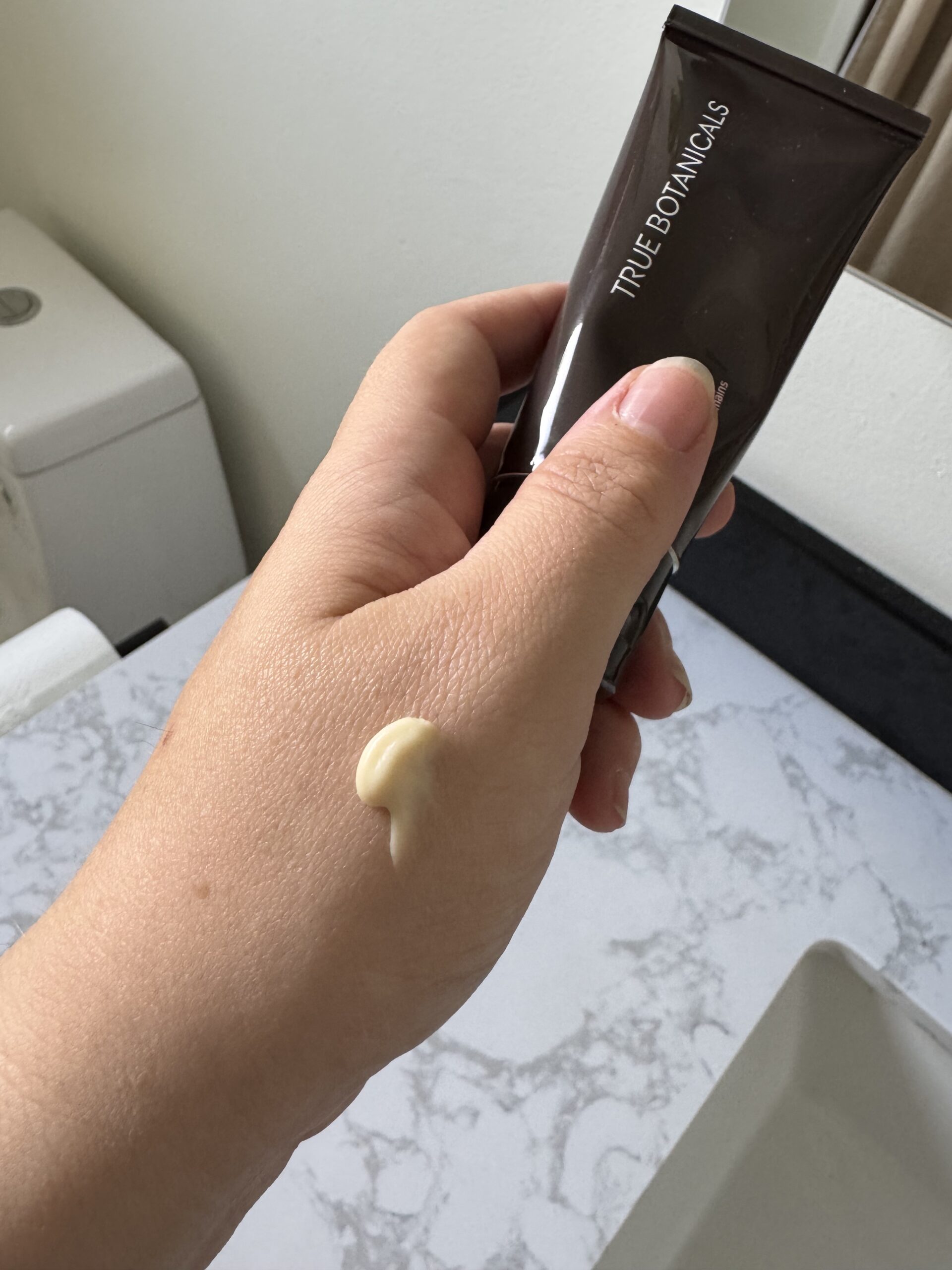 A hand holds a True Botanicals tube with a small amount of cream dispensed onto the back of the hand, in a bathroom setting.