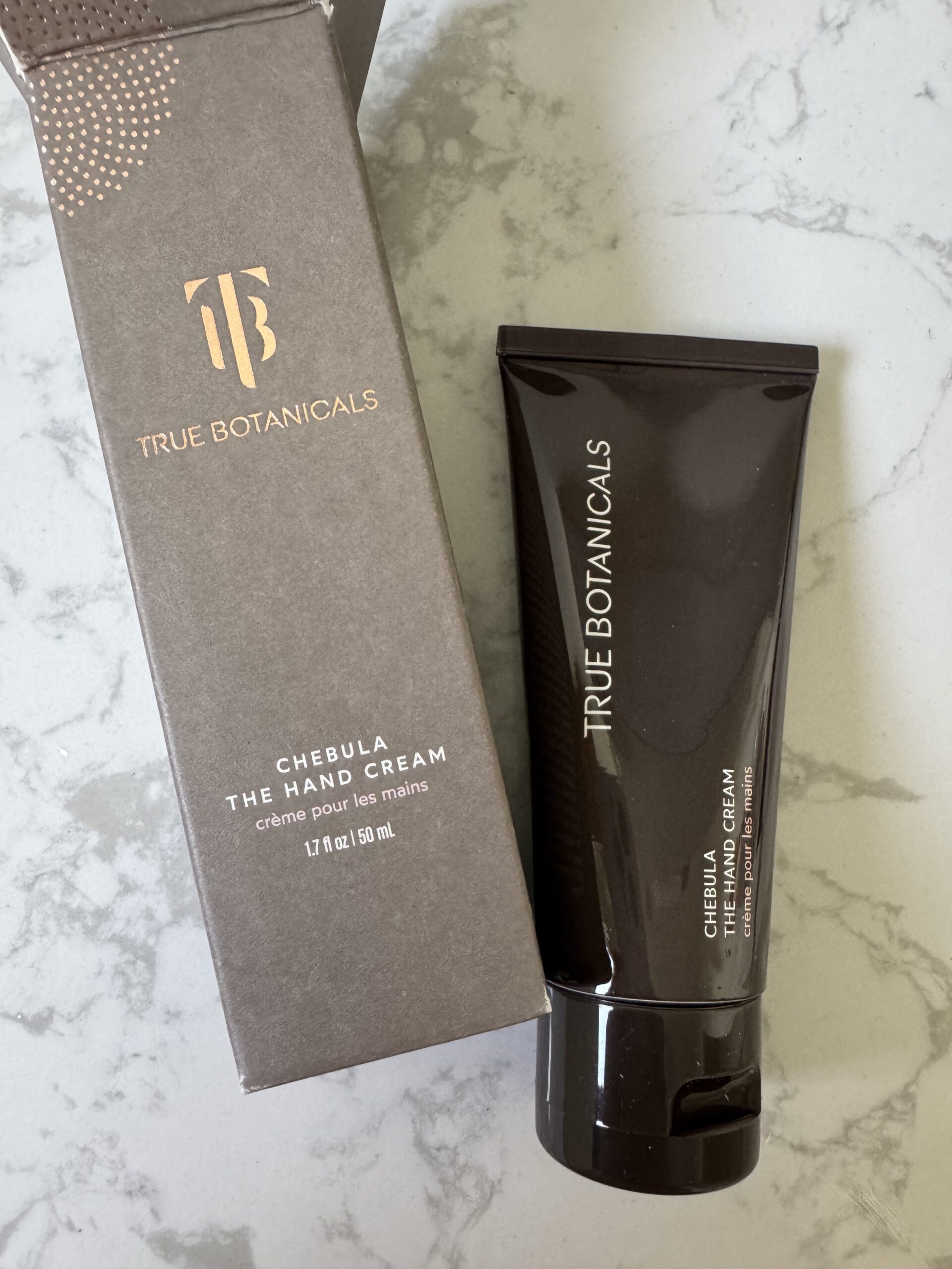 True Botanicals Chebula The Hand Cream tube and its box displayed on a light marble surface.