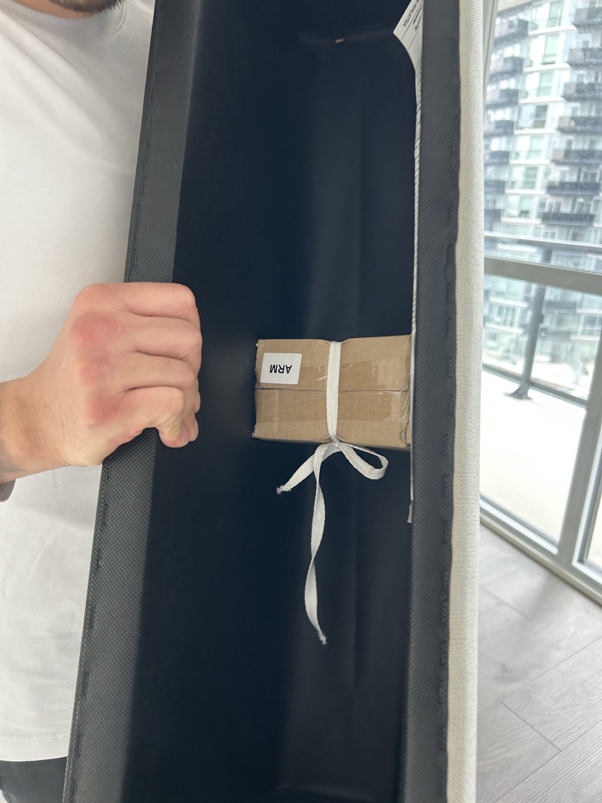 A person holds a large rectangular black container with a small cardboard box tied with white string and labeled "ARM" inside.