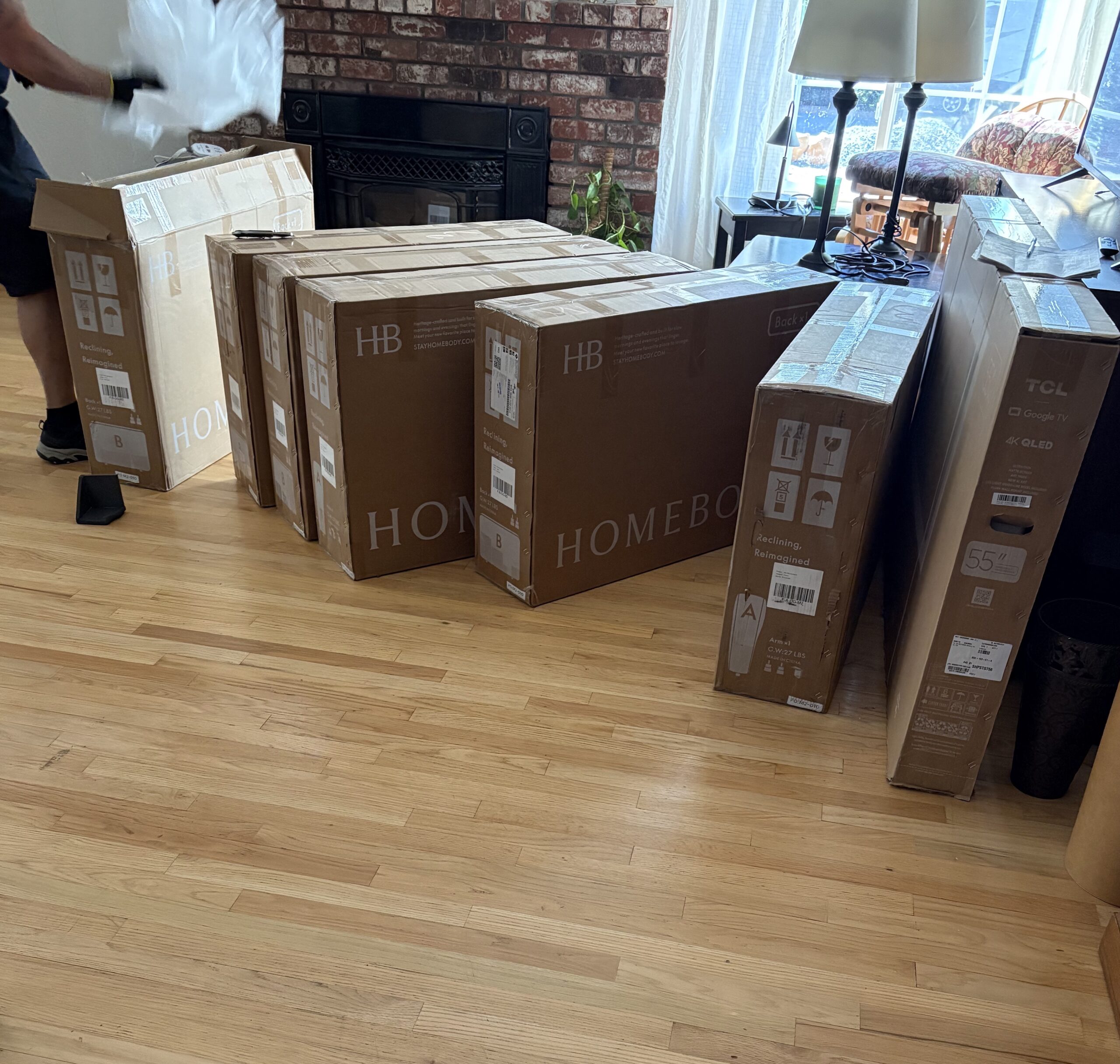 Six large cardboard boxes, including furniture and a TCL 55-inch TV, are arranged on a hardwood floor in a living room. A person’s gloved hand and leg are visible on the left.