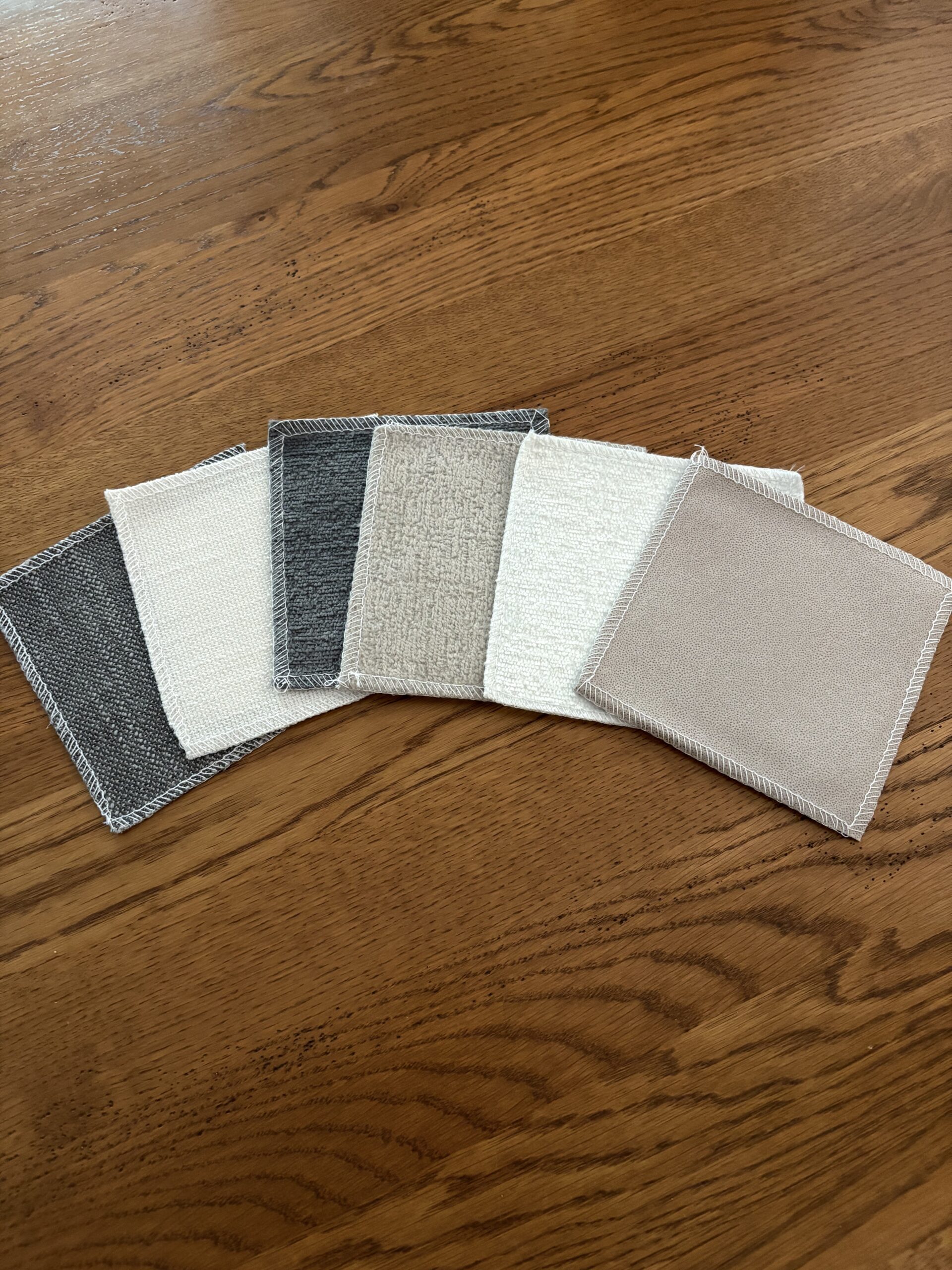 Six square fabric swatches in neutral colors—black, white, gray, cream, taupe, and beige—are arranged in a fan shape on a wooden surface.