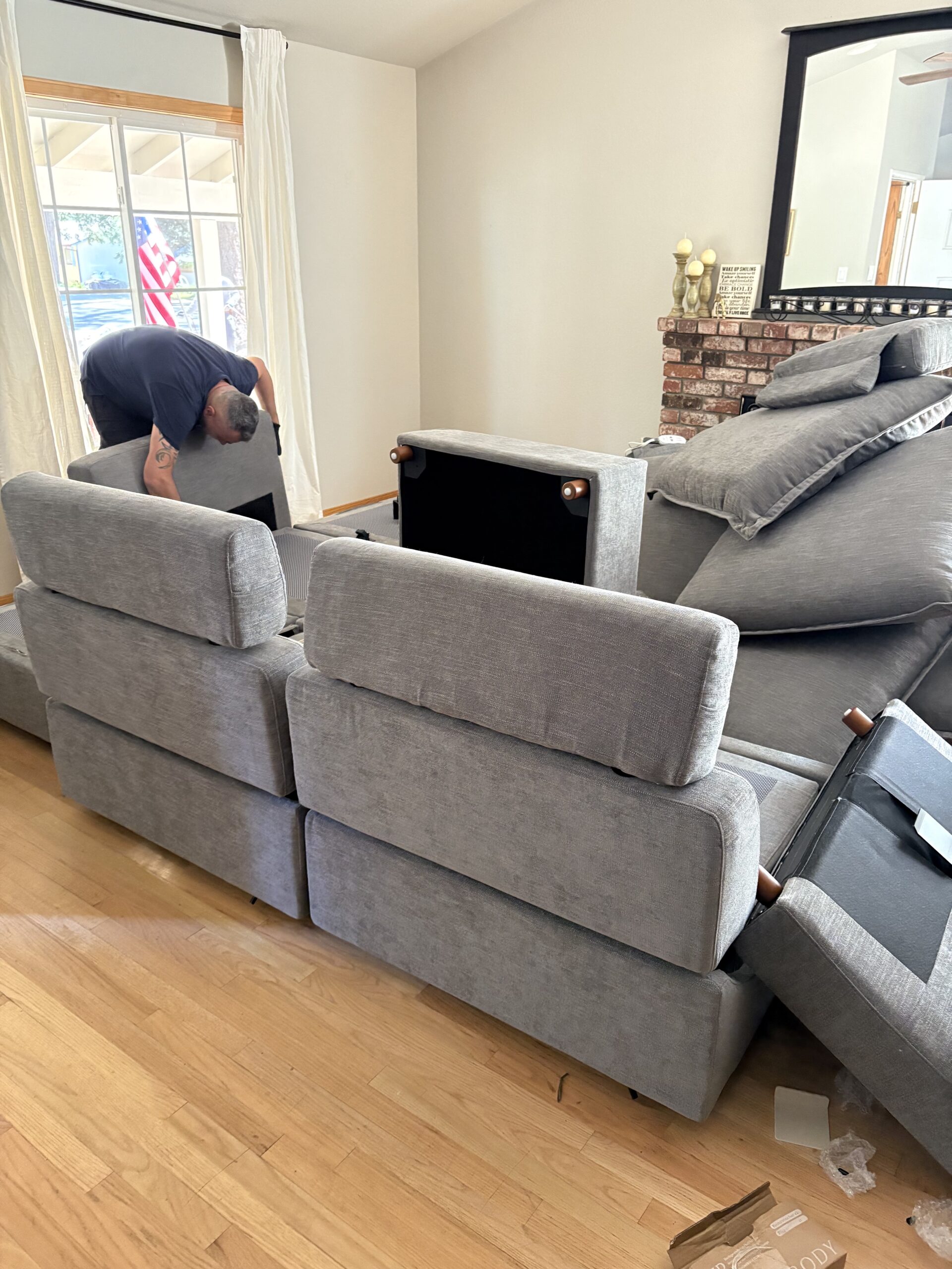 A person is assembling or adjusting a large gray sectional sofa in a living room with hardwood floors and a brick fireplace.