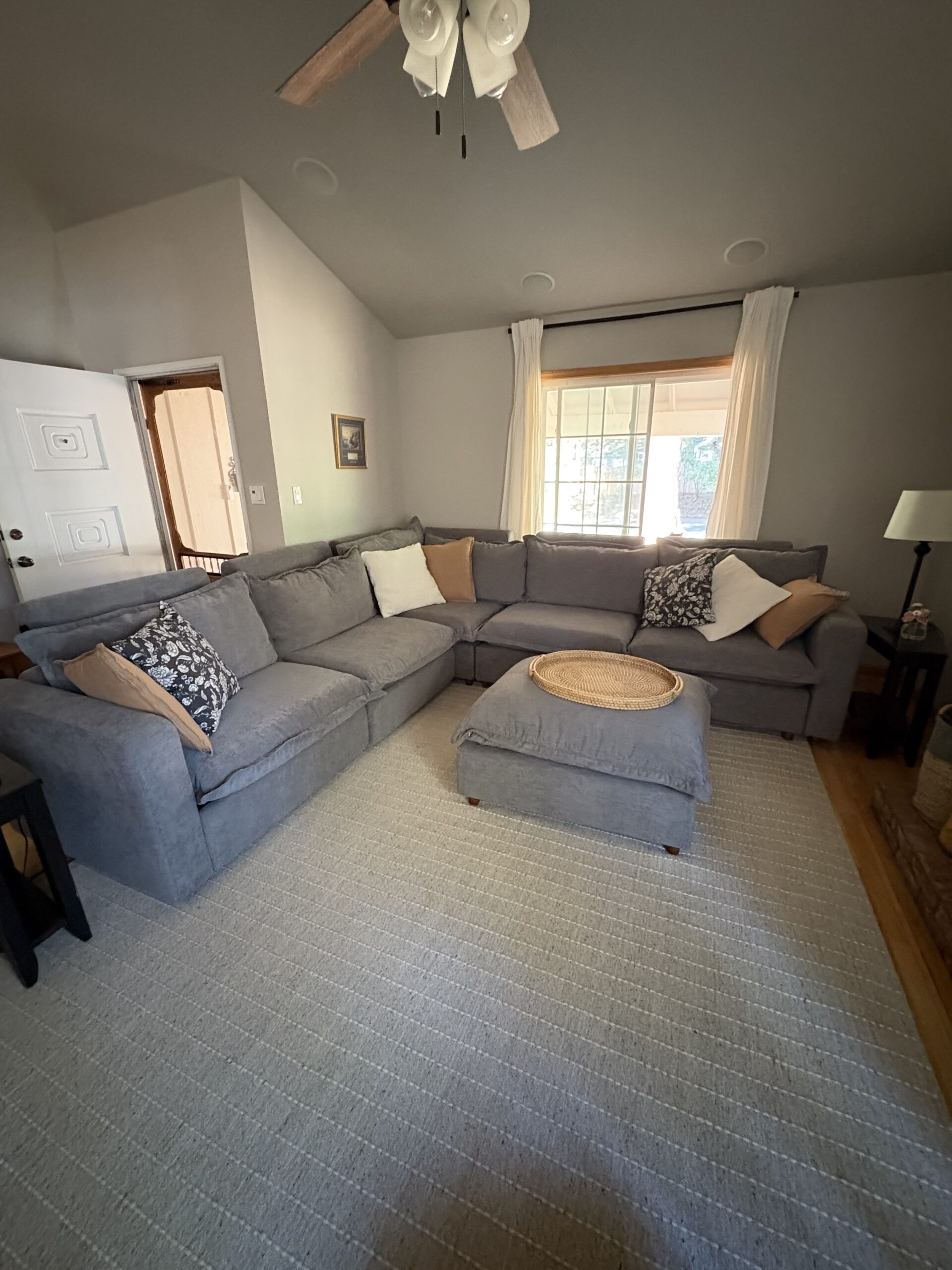 A living room with a large gray sectional sofa, a round tray on an ottoman, several throw pillows, a light rug, a window with white curtains, and a ceiling fan.