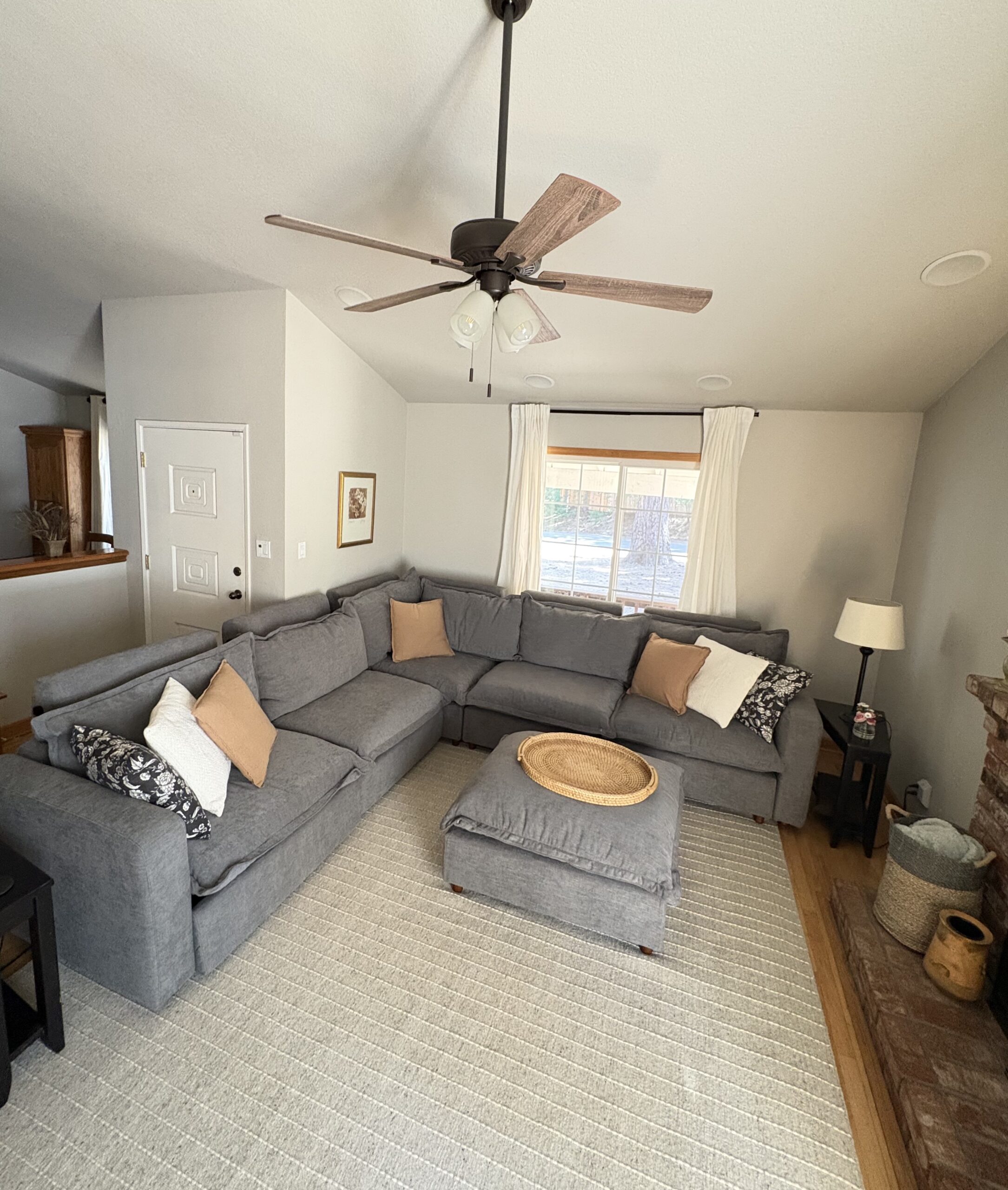 A living room with a gray sectional sofa, beige and patterned pillows, a matching ottoman with a tray, a lamp, and a window with beige curtains. A ceiling fan is centered above.