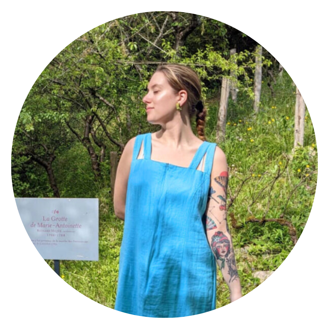 A woman in a blue sleeveless dress stands outdoors near a sign, with greenery and trees in the background. She has tattoos on her left arm and is looking to the side, appearing relaxed as if enjoying the comfort of a latex mattress.